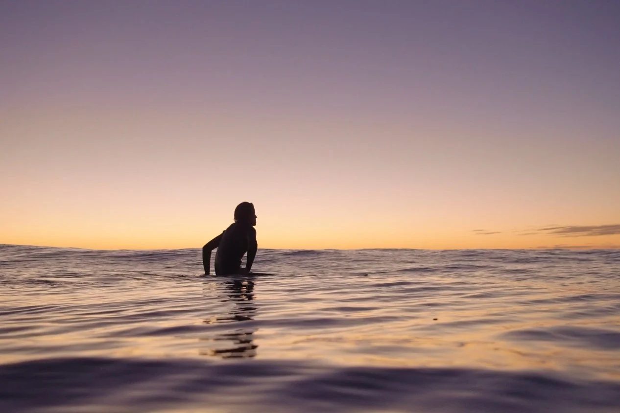 Silhouette of a person sitting in the ocean at sunset or sunrise with a colorful sky.