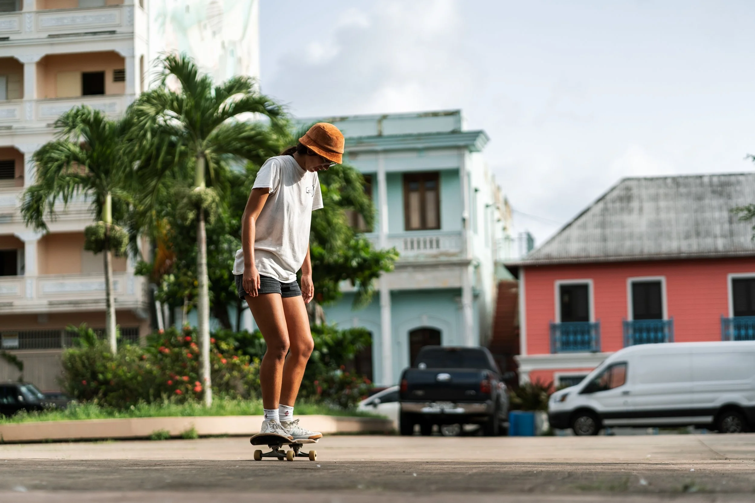 A young woman skateboarding outdoors on a city street with colorful buildings and palm trees in the background.