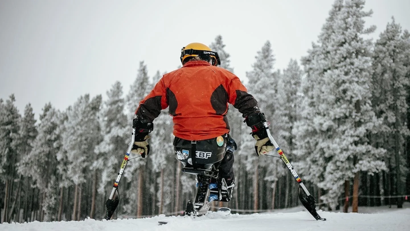 A person in a bright orange and black winter jacket with a yellow helmet, using snow skis and poles, standing on snow with a forest of snow-covered trees in the background.