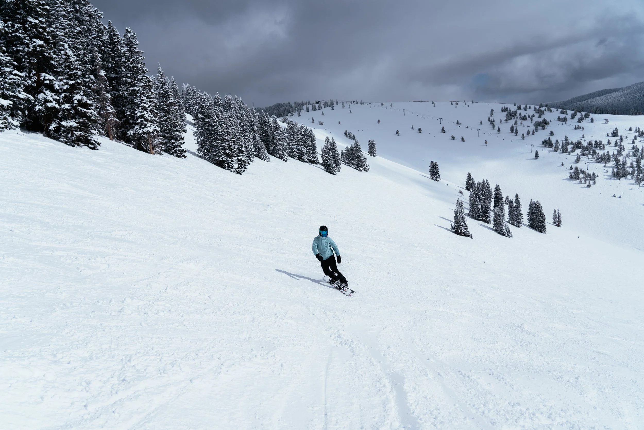 A person snowboarding on a snowy mountain slope with pine trees, dark clouds overhead, and distant rolling hills.