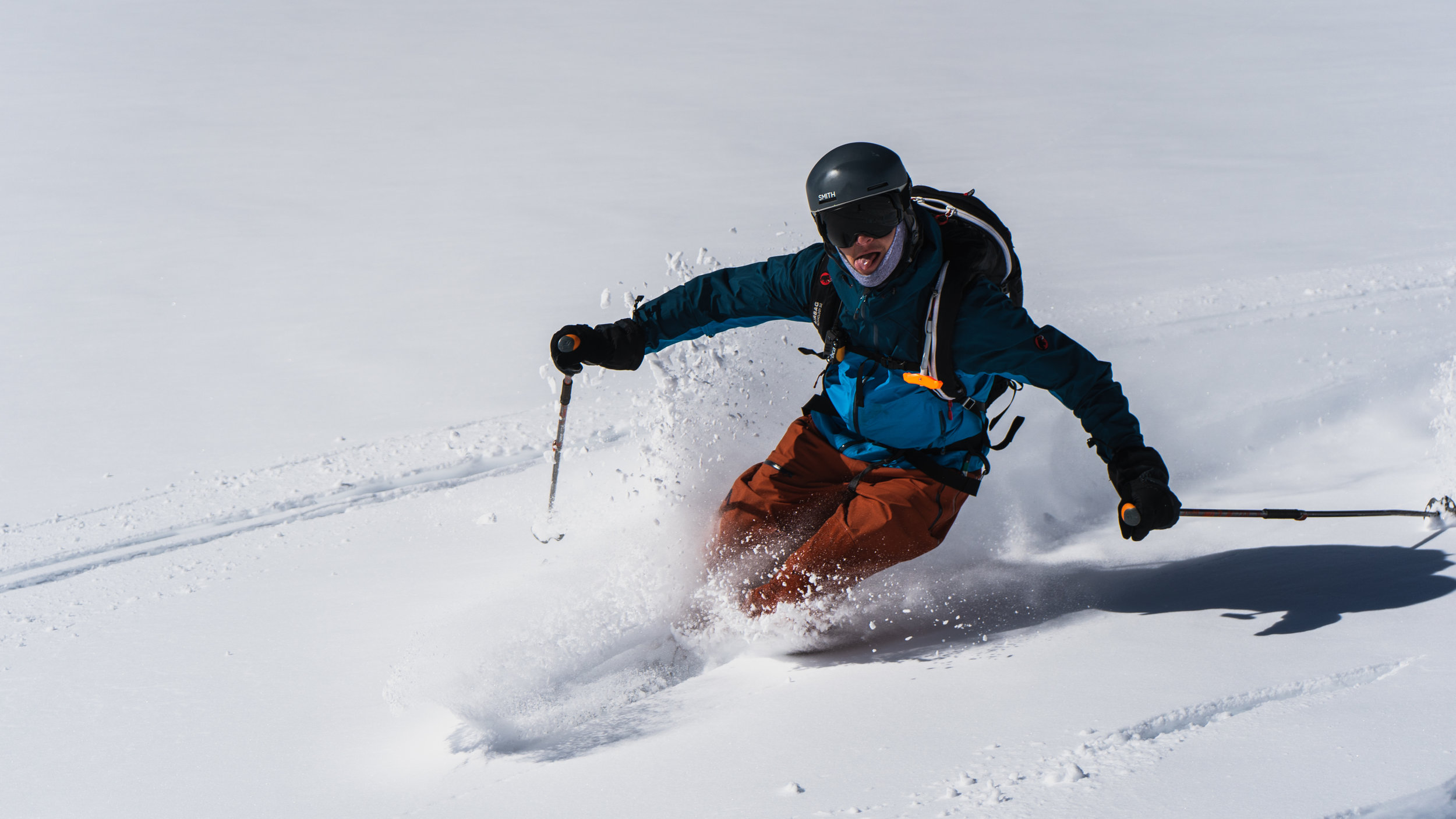 A person skiing in the snow, wearing a black helmet, goggles, a blue jacket, and orange pants, creating a spray of snow as they descend down a slope.