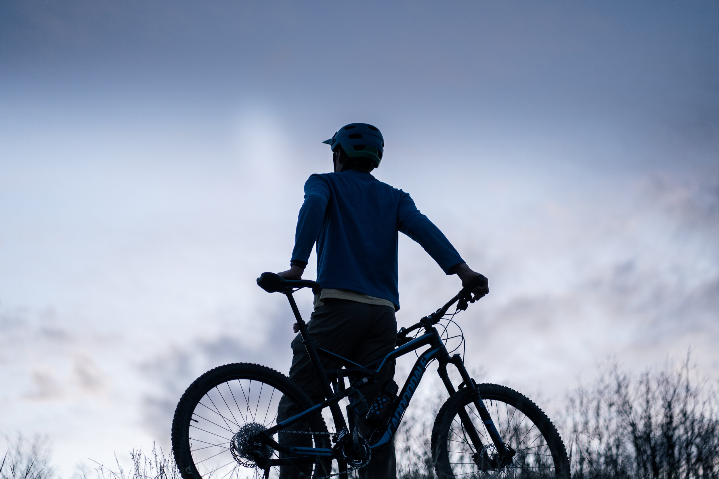 A person wearing a helmet, standing with a mountain bike against a cloudy sky.