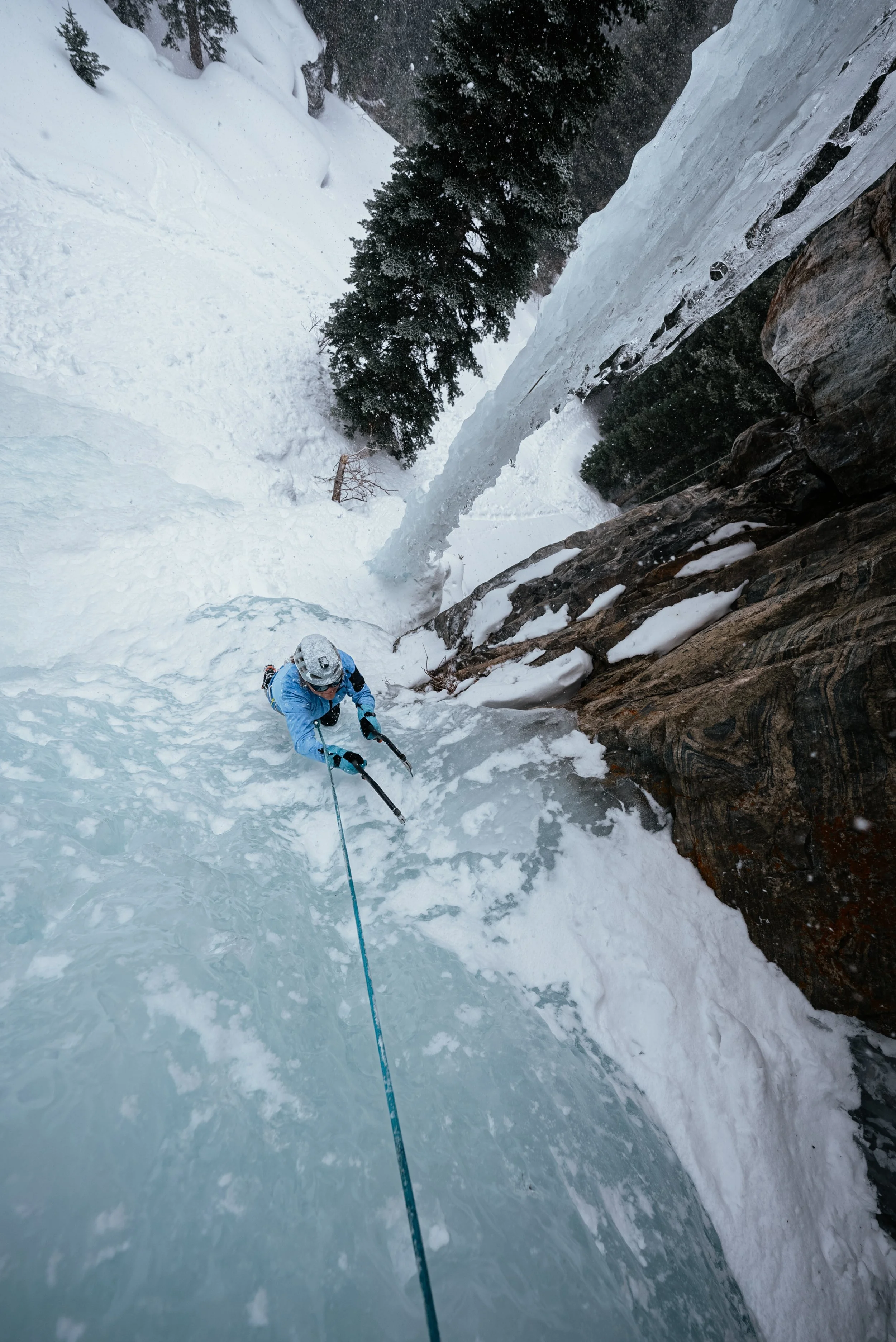 A person in blue outdoor gear and a helmet is climbing a frozen waterfall with ice axes, secured by a safety rope, in a snowy landscape with trees and rocks.