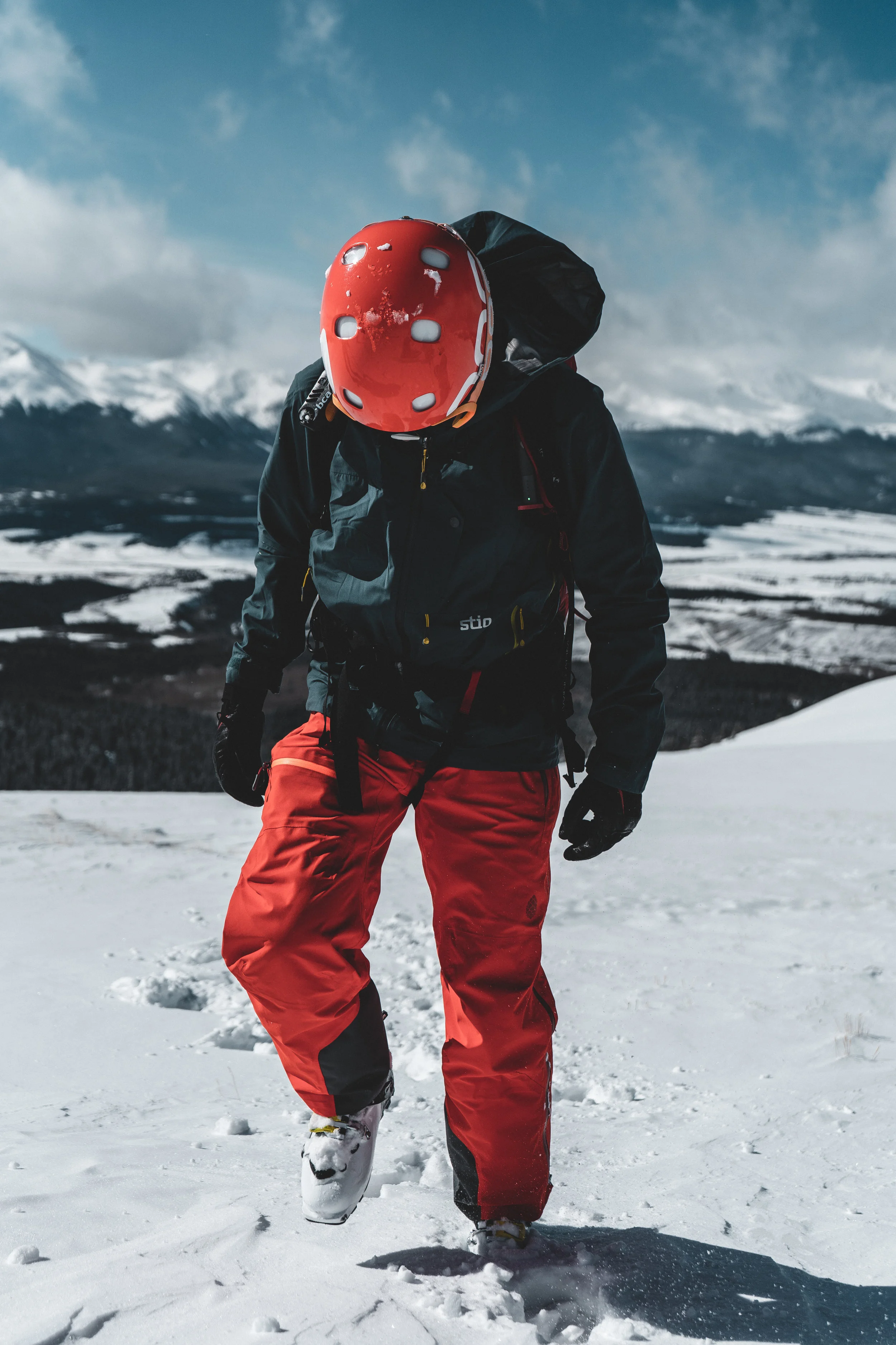 Person dressed in winter gear with a red helmet and red snow pants walking in snowy terrain with mountains in the background.