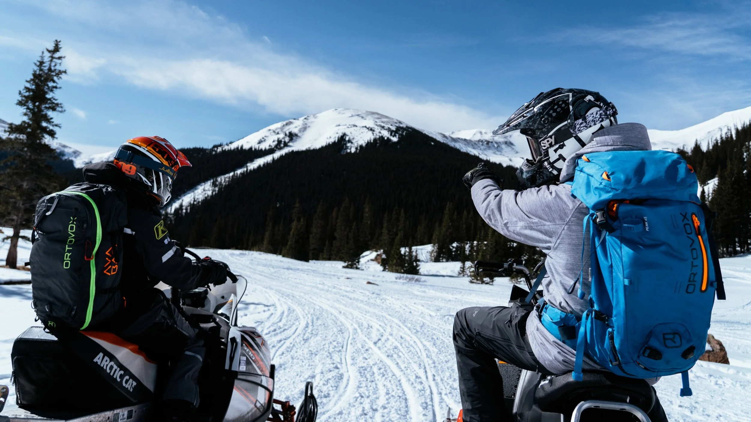 Two people in snow gear riding snowmobiles in a snowy mountain landscape with pine trees and a clear blue sky.