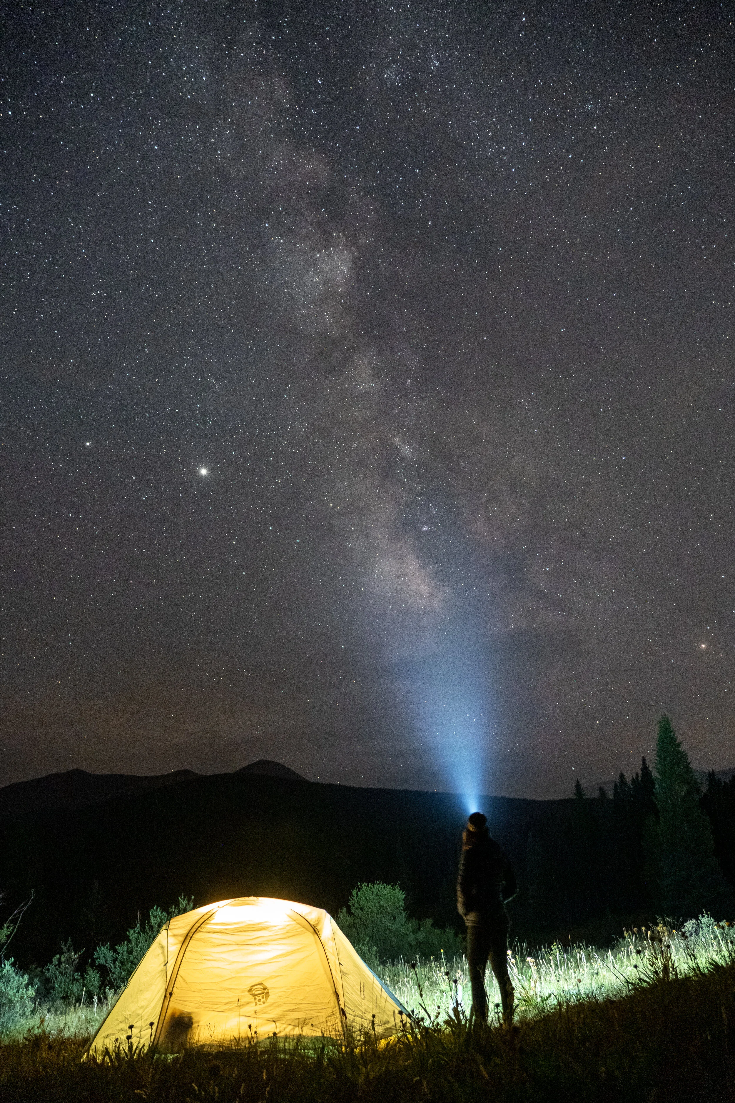 A person camping outdoors at night under a star-filled sky with the Milky Way galaxy visible, a yellow tent illuminated from within, and the person shining a flashlight or headlamp towards the sky.