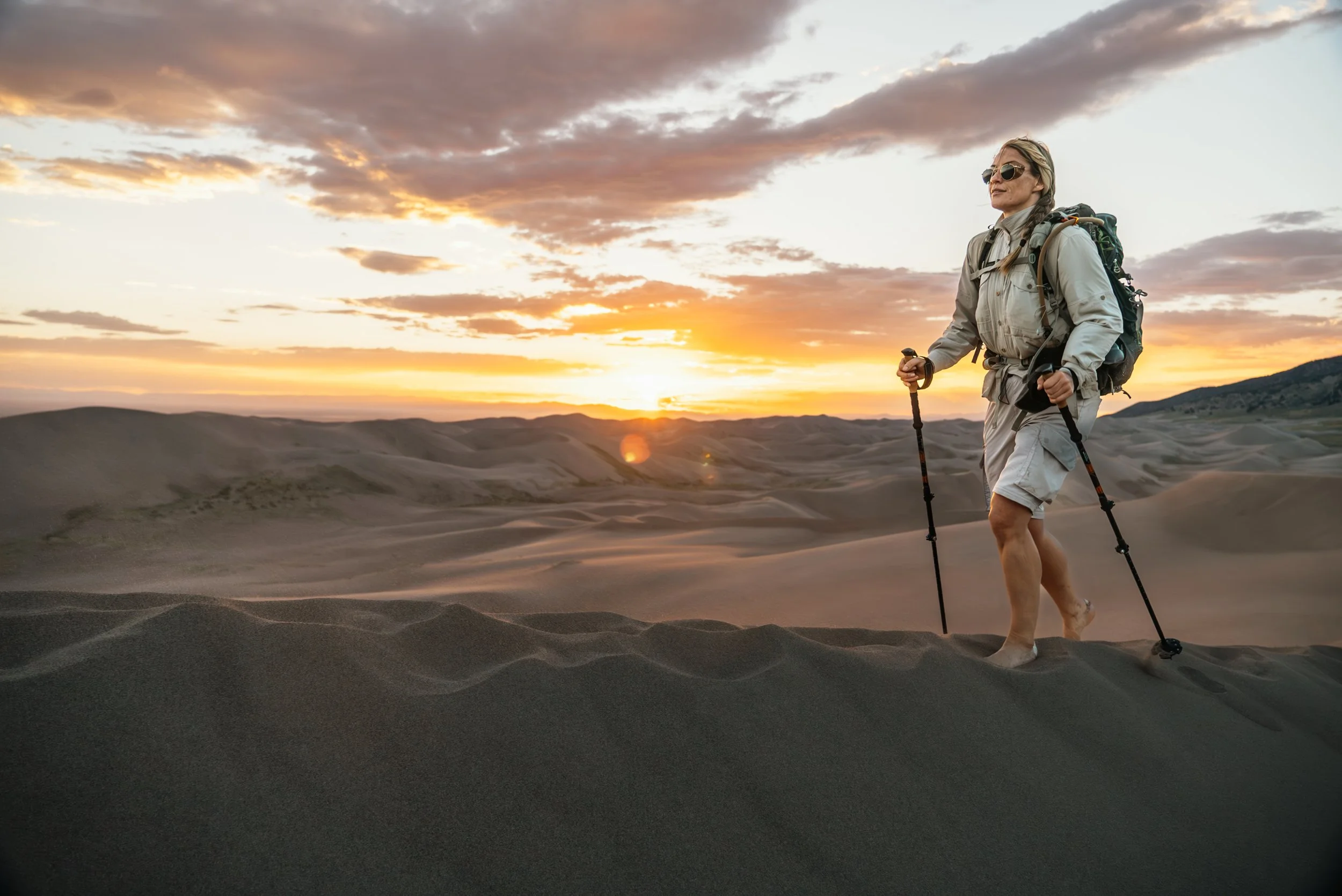 A woman hiking in the desert at sunset, carrying a backpack and using trekking poles.