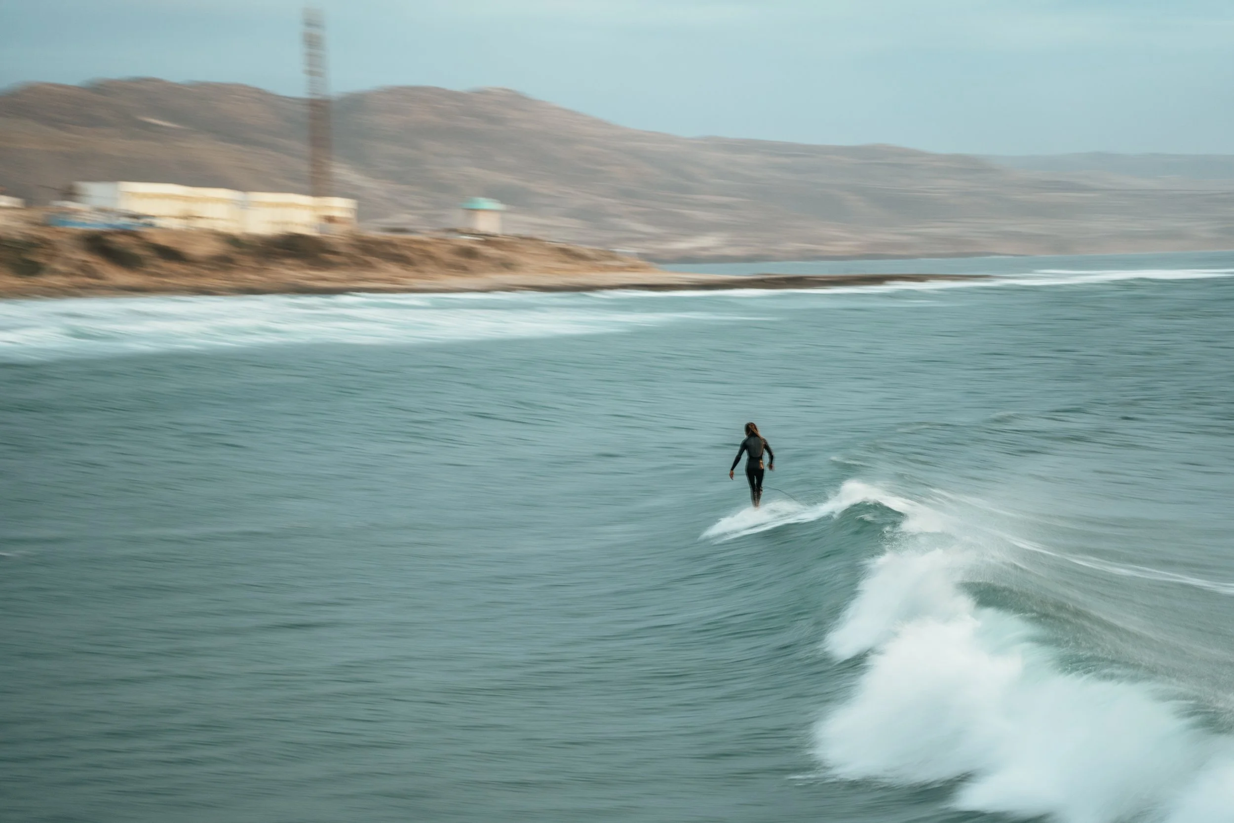 A person surfing on a wave in the ocean with a hilly landscape and some buildings in the background.
