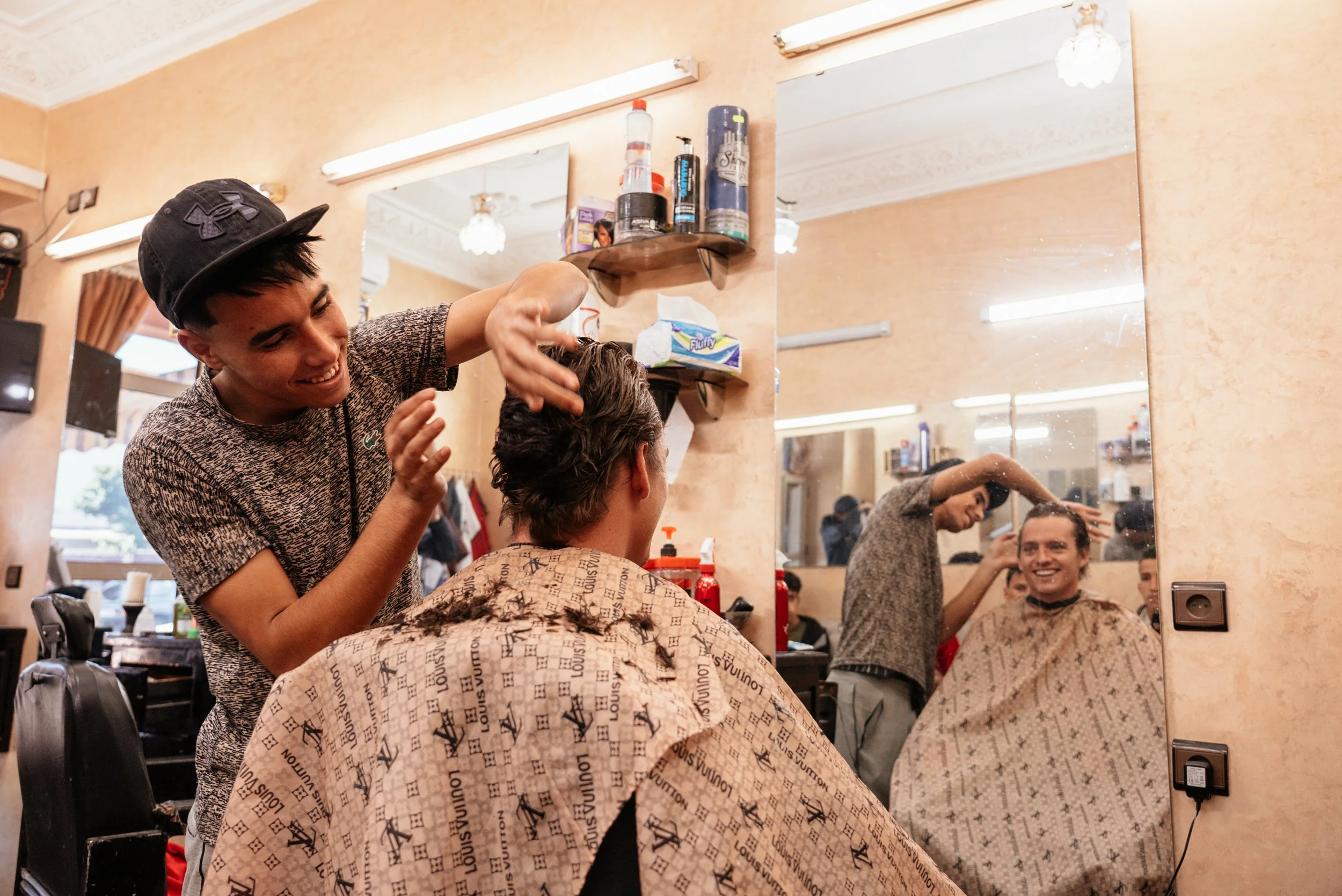 A young man with dark hair receiving a haircut in a salon, smiling at his reflection in a large mirror.