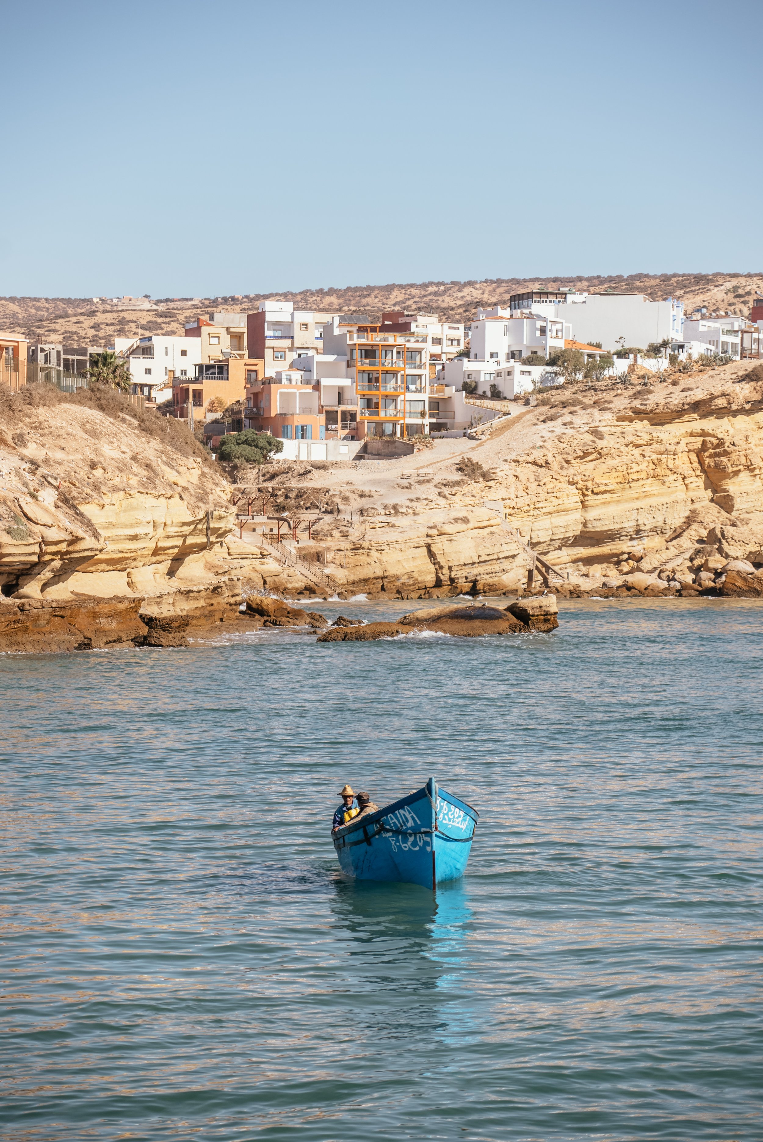 A small blue boat floating in calm water near a rocky shoreline with multi-story residential buildings on a hillside in the background.