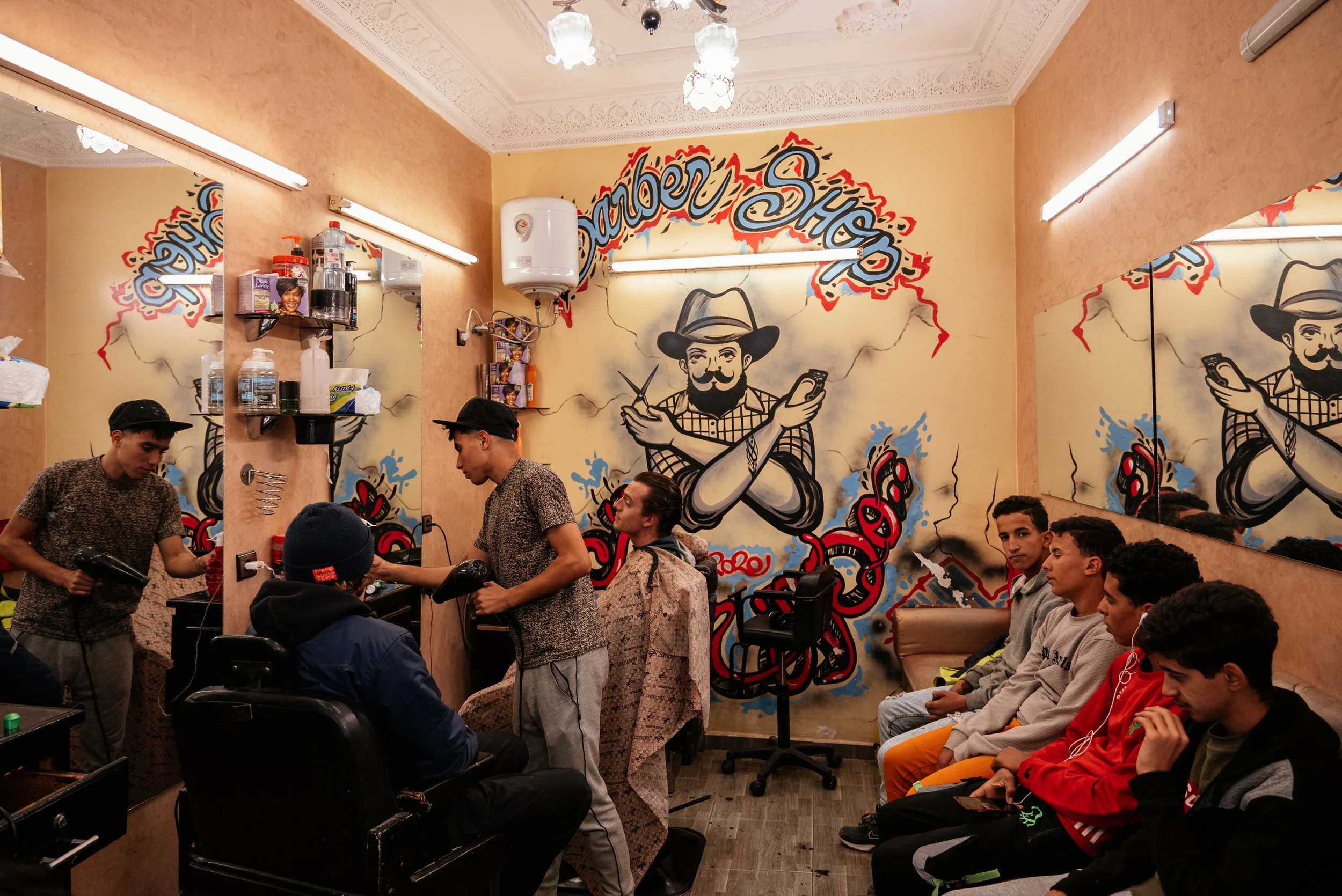 Barbershop with a mural of a man with crossed arms holding scissors and a comb on the wall. Several young men are waiting, and two barbers are cutting a customer's hair.