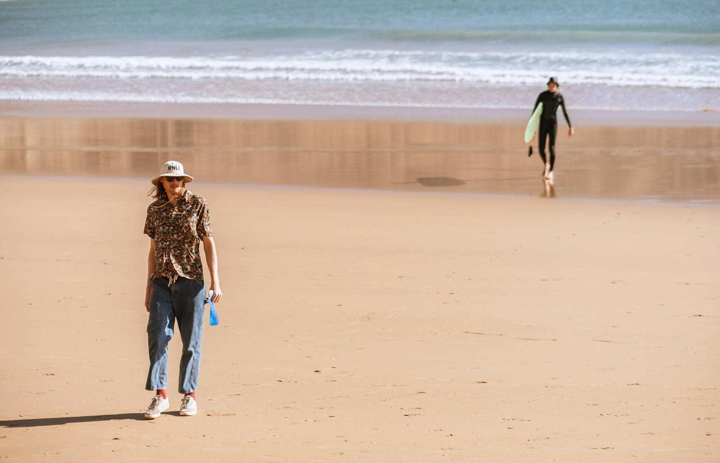 A woman walking on a sandy beach holding a water bottle, wearing a hat, sunglasses, and casual clothing. In the background, a person is walking with a surfboard toward the water.