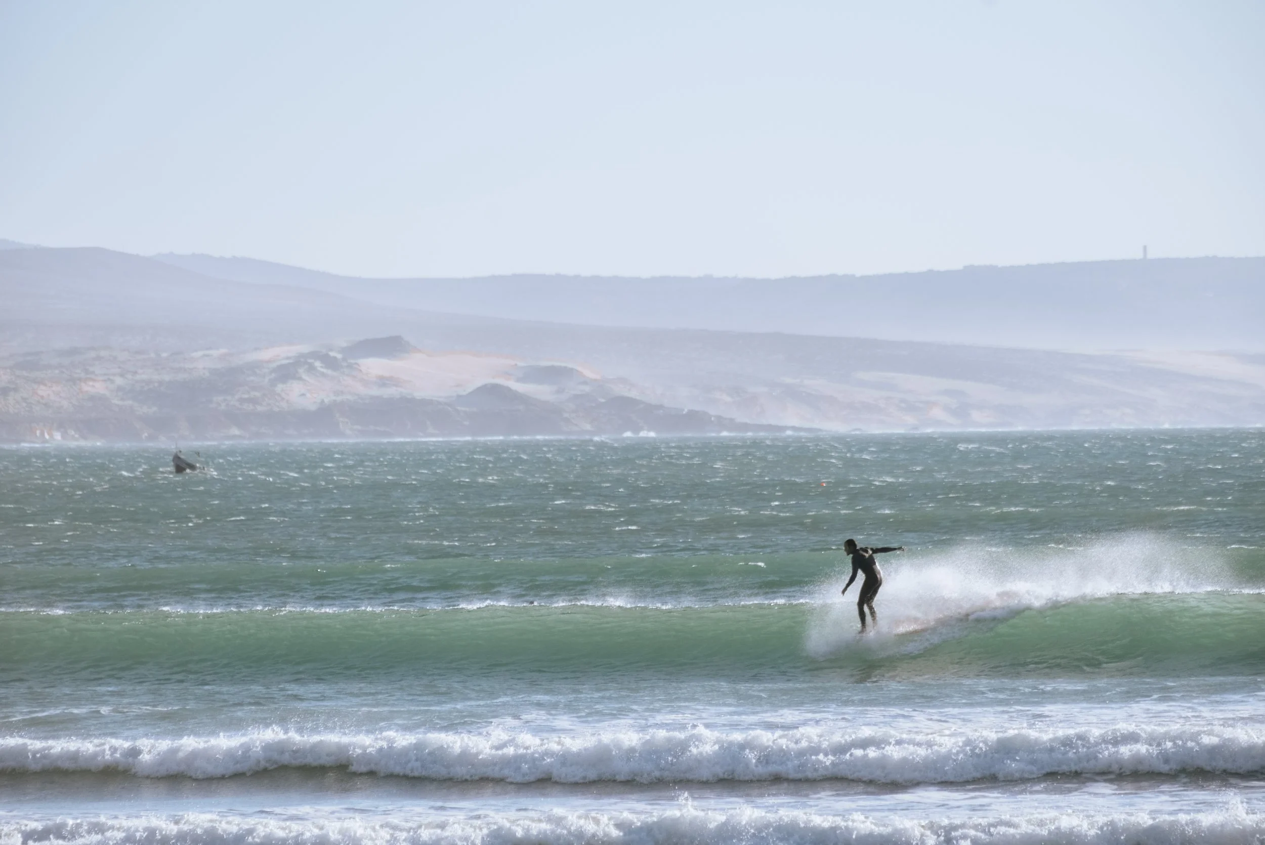 A person surfing on a wave near the shore with a boat in the background and hilly landscape in the distance.