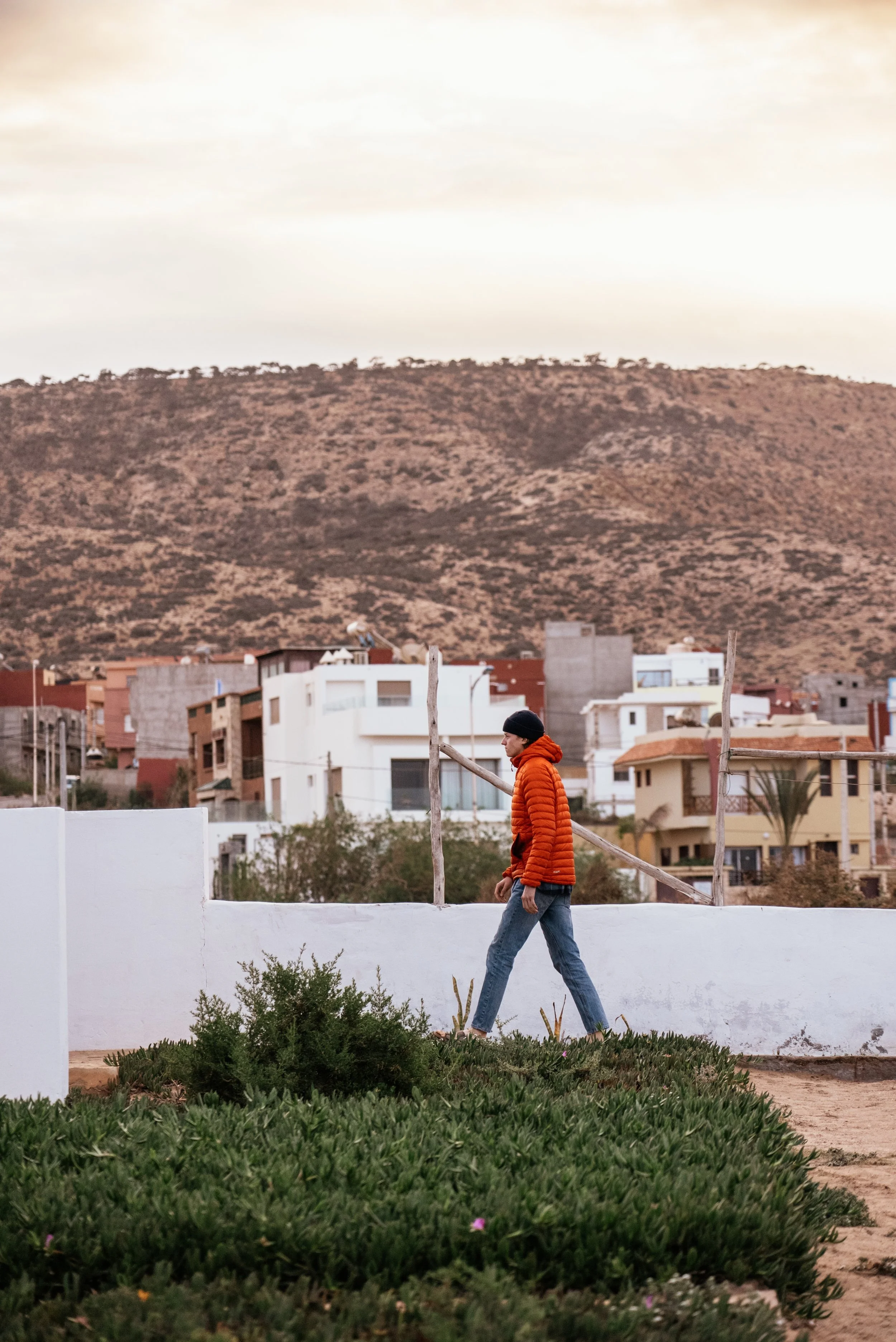 A man in a red puffer jacket, jeans, and a black beanie walking on a path with green plants in the foreground, against a background of colorful houses and a mountain under a cloudy sky.