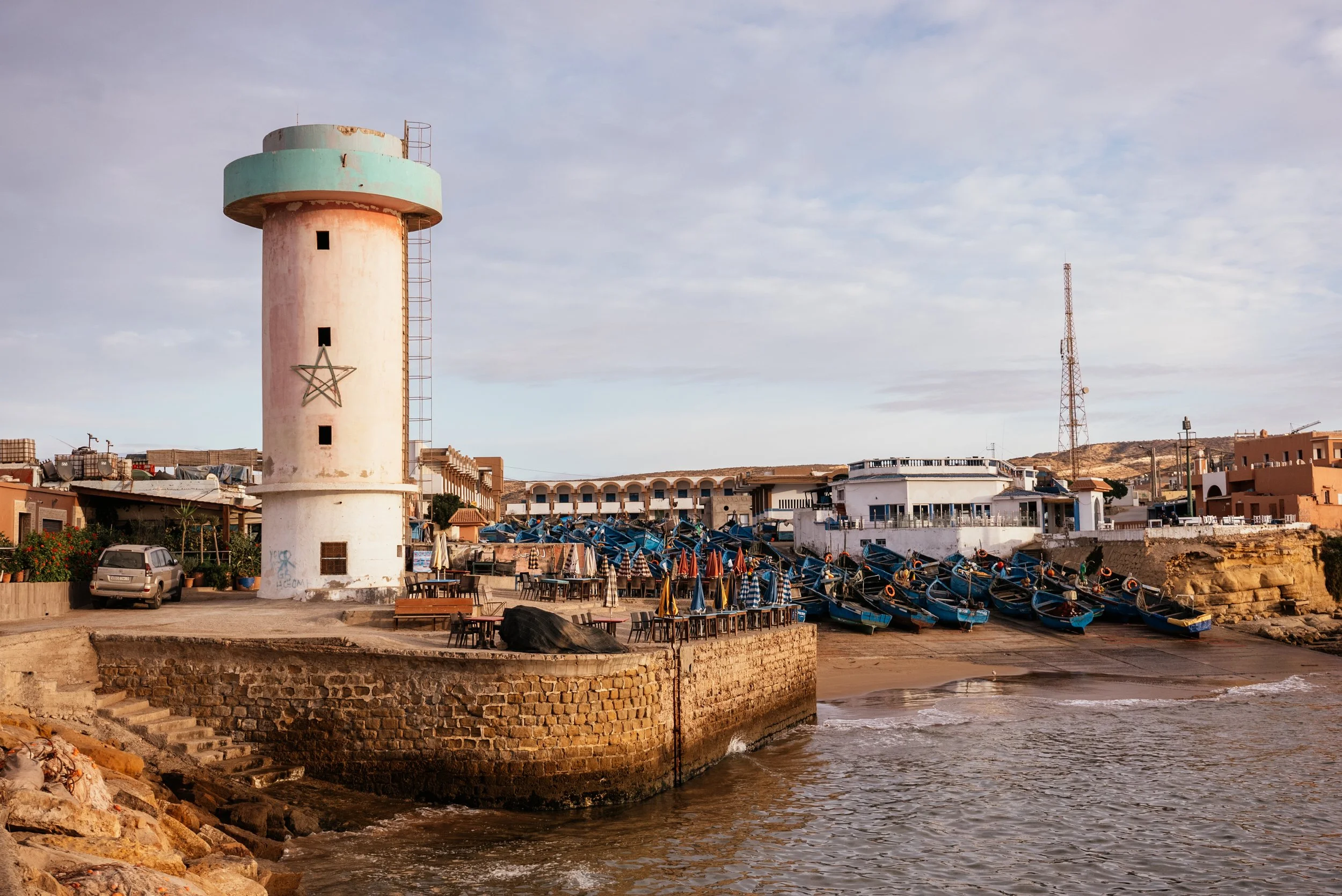A coastal scene with a white lighthouse featuring a star symbol on its side, several boats pulled up on the beach, and surrounding buildings under a partly cloudy sky.