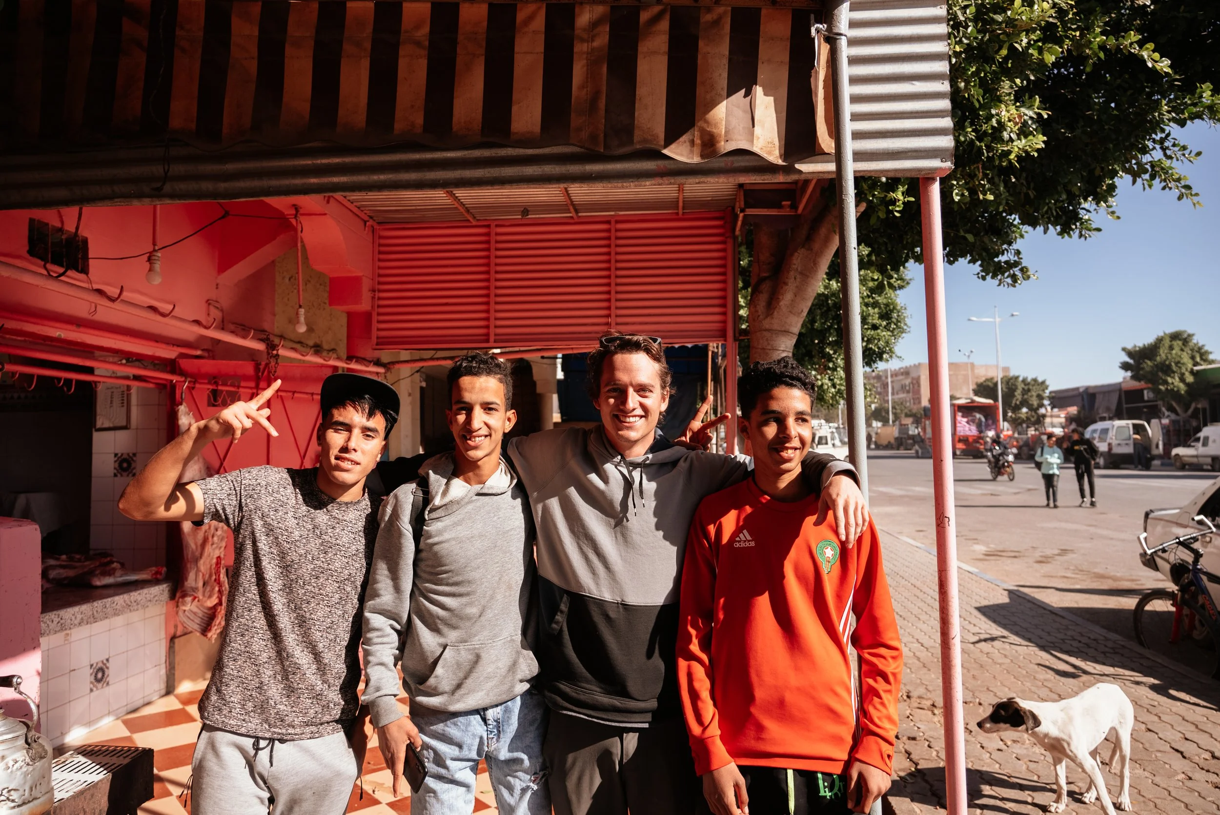 Four young men standing together outside a building, smiling and posing for the photo. One man is wearing a gray hoodie, another a red sports jacket, and the others casual shirts. A dog is standing nearby on the sidewalk, and there are trees, cars, a