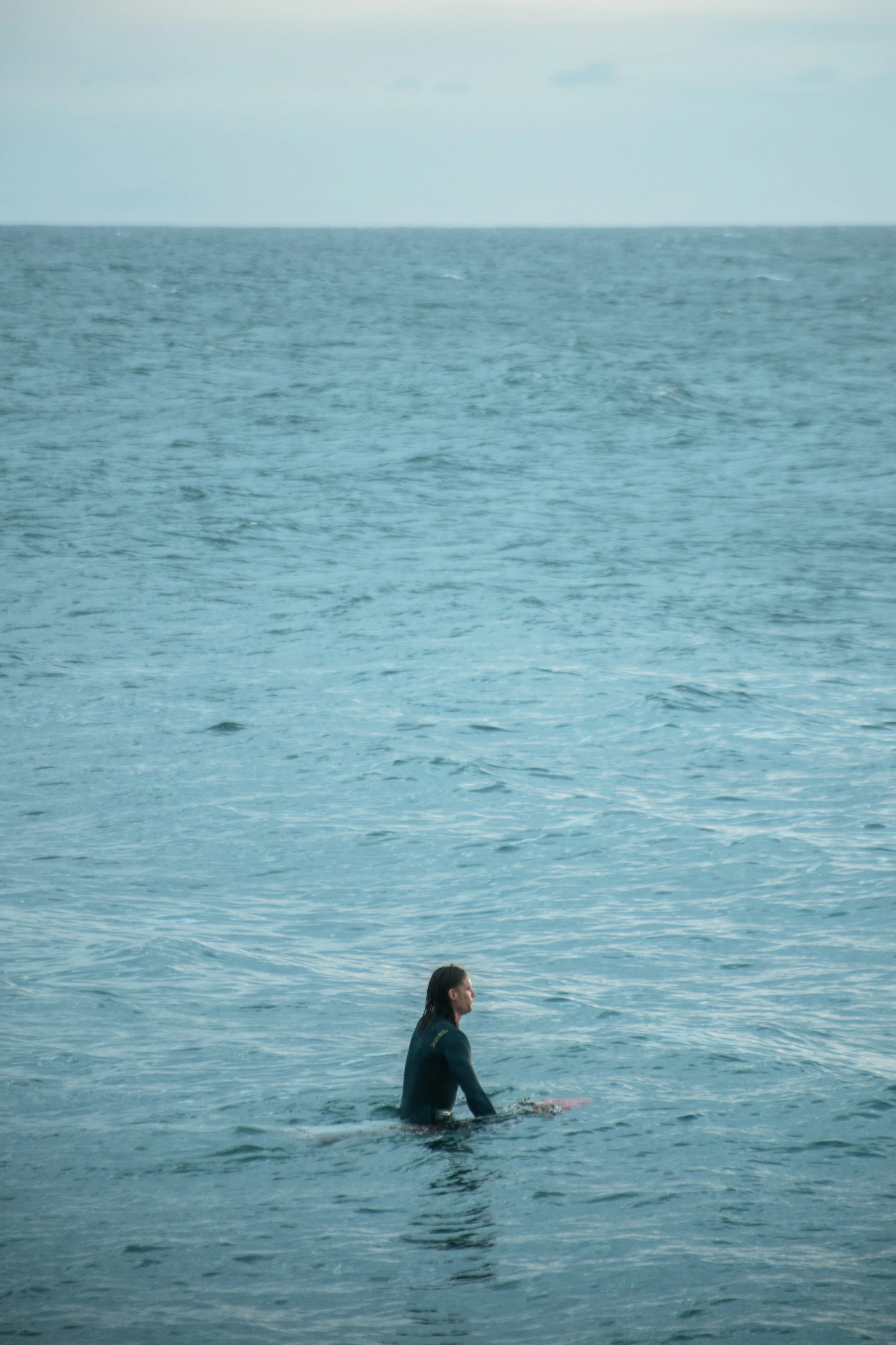 Person in a wetsuit sitting on a surfboard in the ocean, with the water stretching out to the horizon under a cloudy sky.