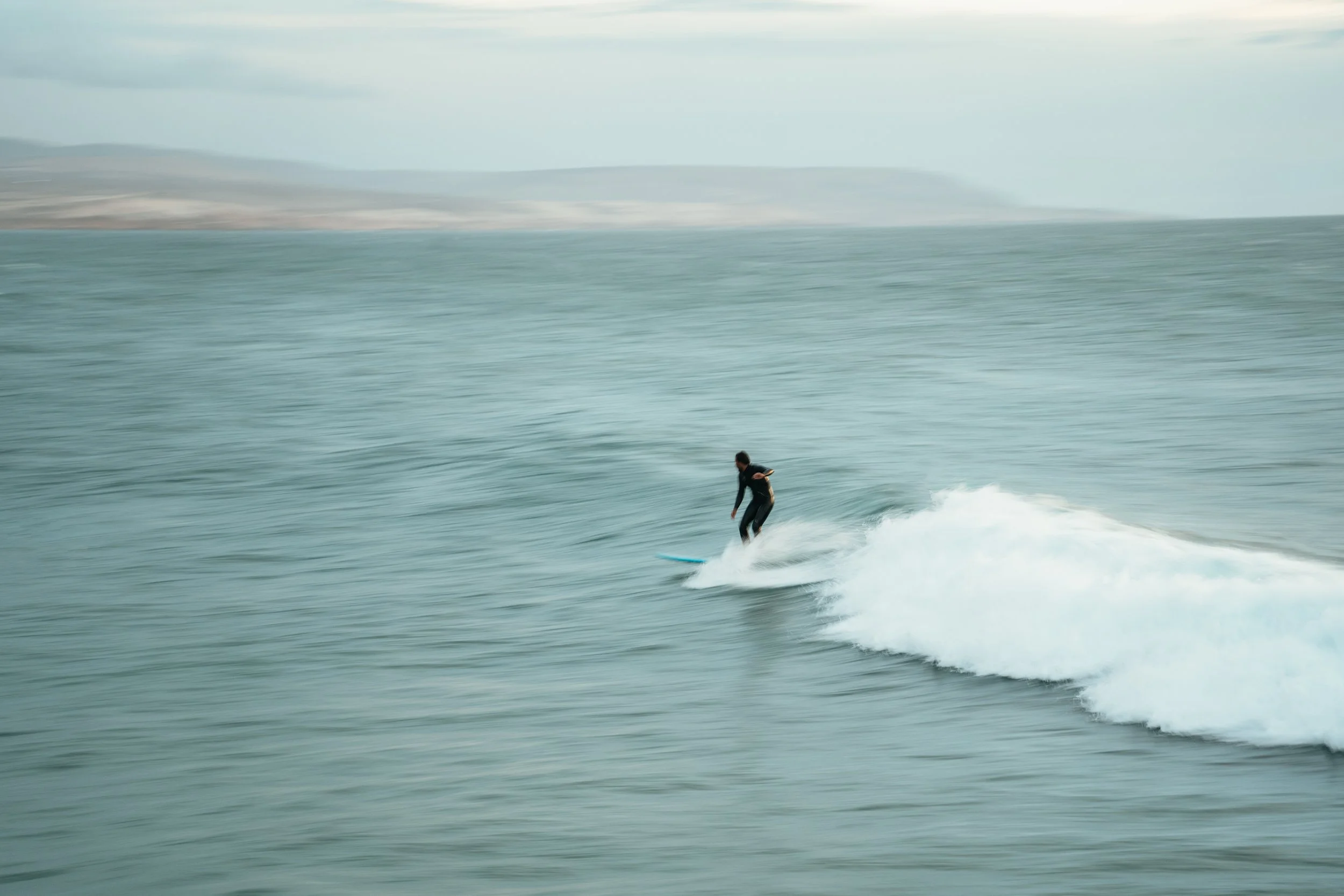 A person surfing on a wave in the ocean, with distant land and cloudy sky in the background.