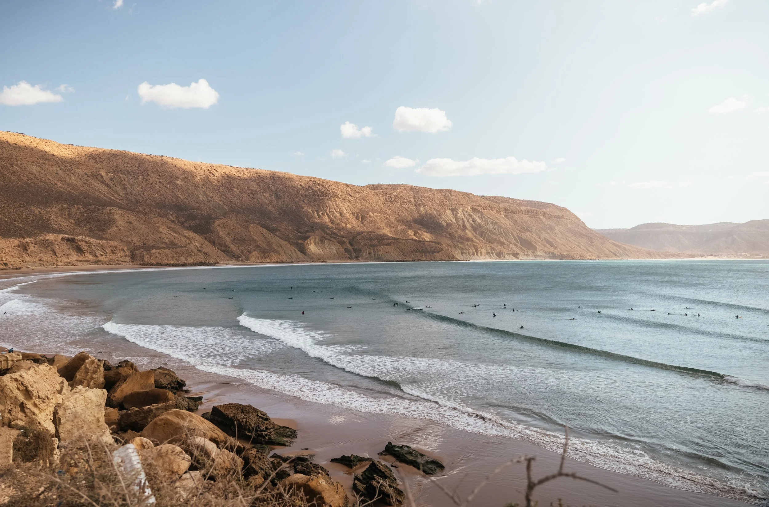 Beach with rocks in the foreground, gentle waves, surfers on the water, and hills in the background under a partly cloudy sky.