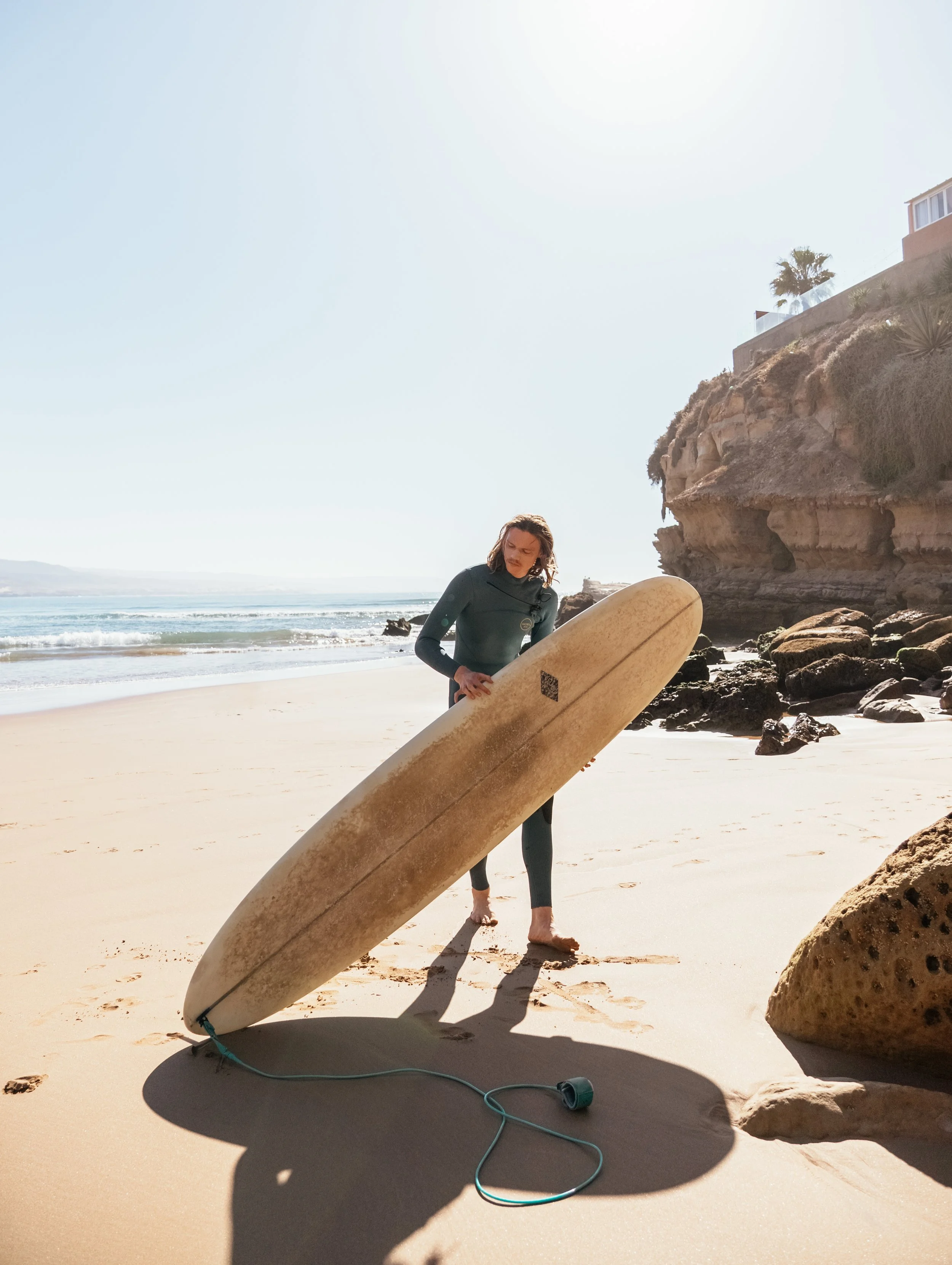 A person holding a surfboard on the beach, preparing for surfing, with rocks and cliffs in the background under a bright sun.