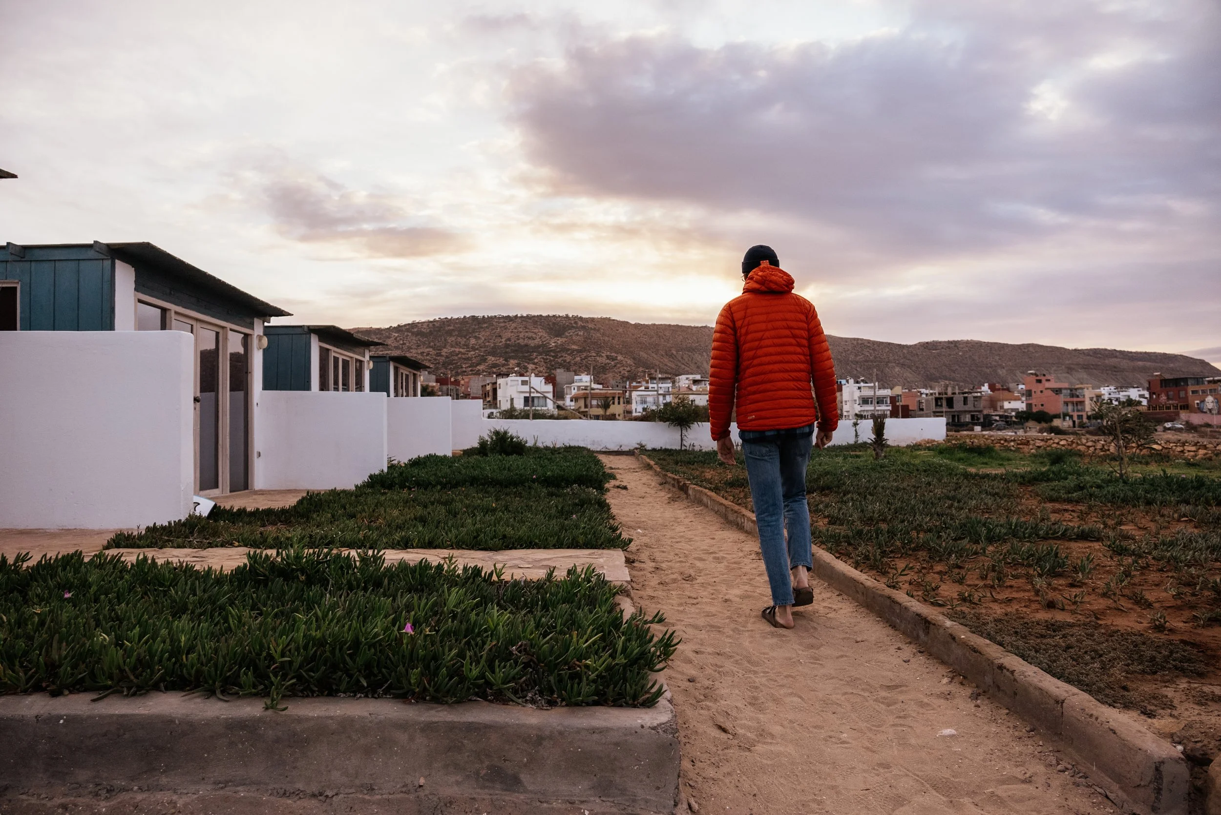 A person dressed in an orange jacket, jeans, and sandals walking along a dirt path in a residential area with white houses and greenery, with hills and a cloudy sky in the background.