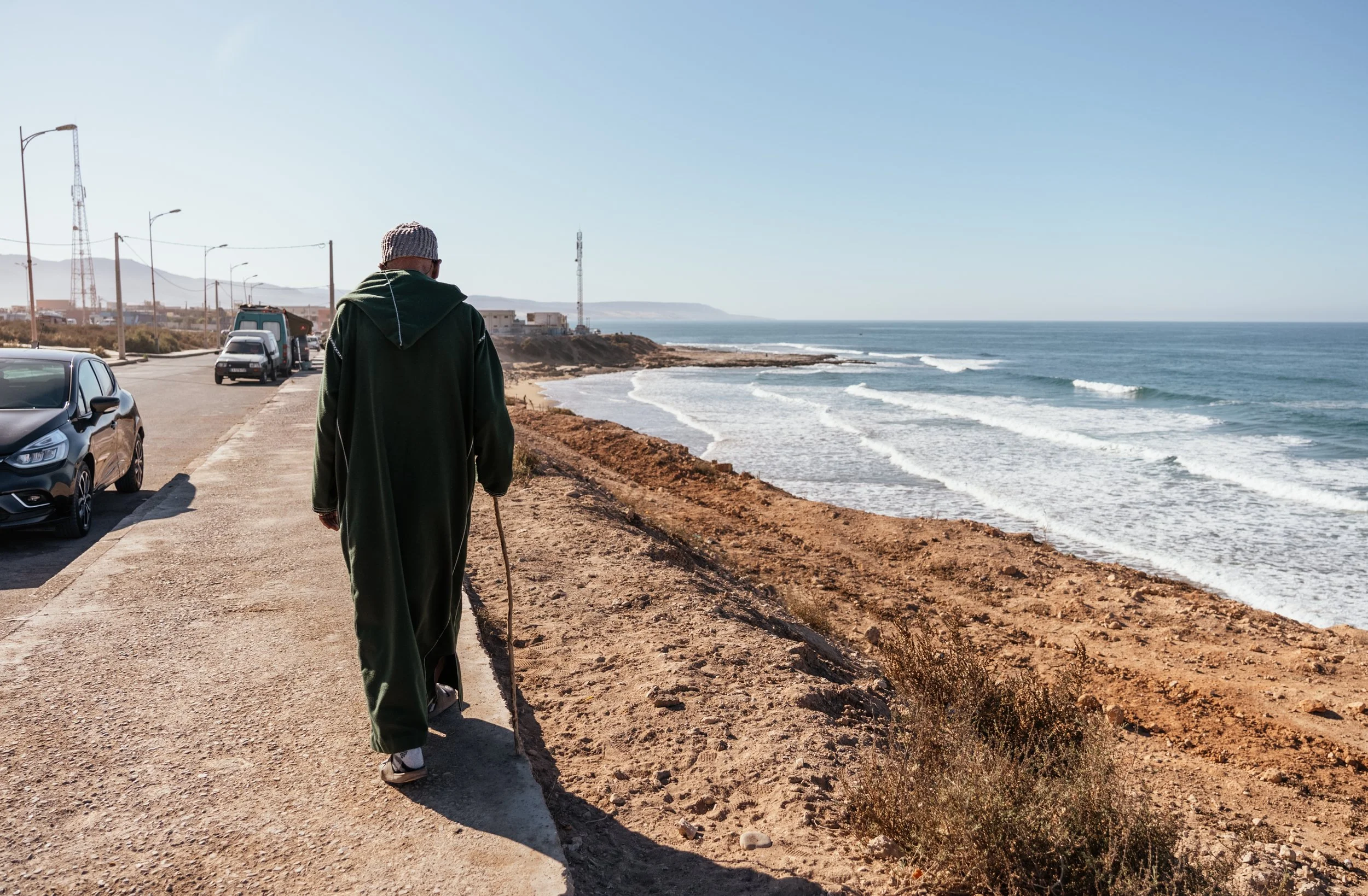 A person walking along a coastal road with cars parked nearby, overlooking the ocean with waves, and distant buildings and hills under a clear blue sky.