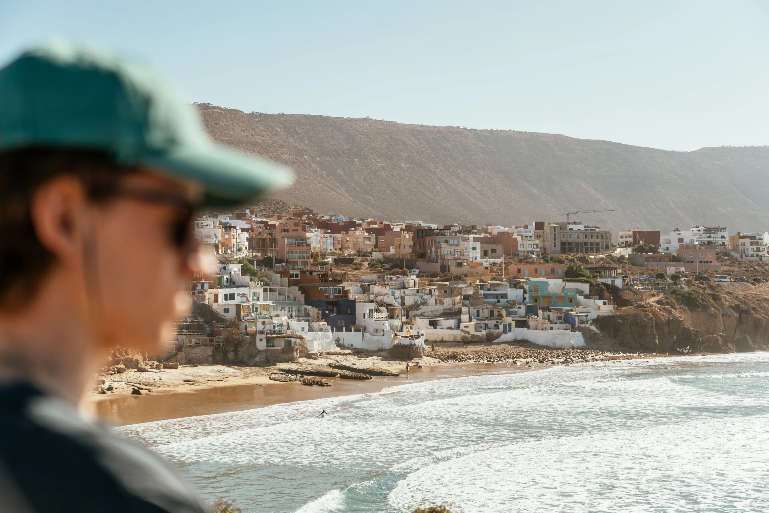 Blurred person wearing a green cap and sunglasses in foreground, overlooking a beach with colorful houses on a hillside and ocean waves in the background.