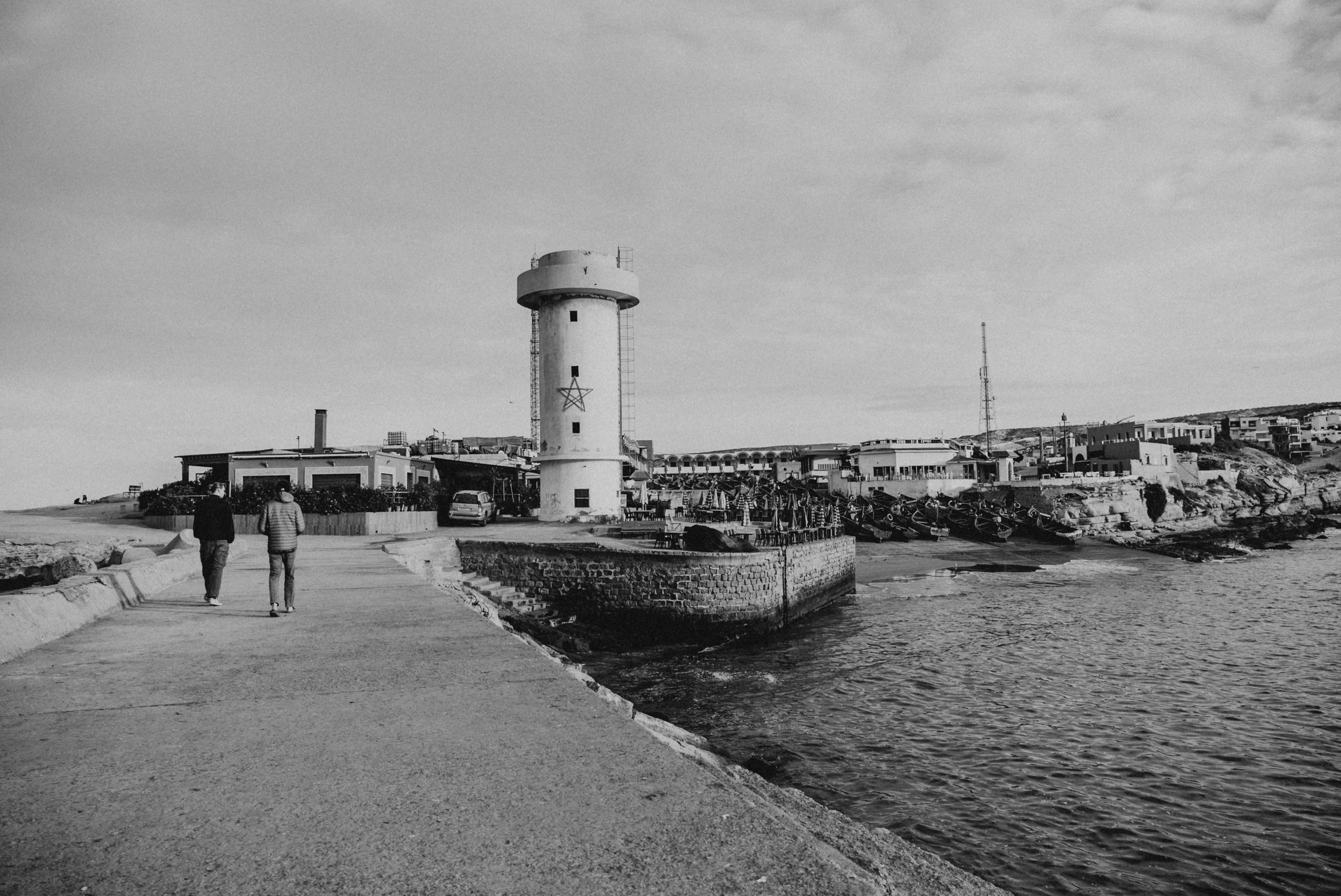 Black and white photo of a seaside promenade with two people walking toward a lighthouse, boats docked along the shoreline, and buildings in the background.