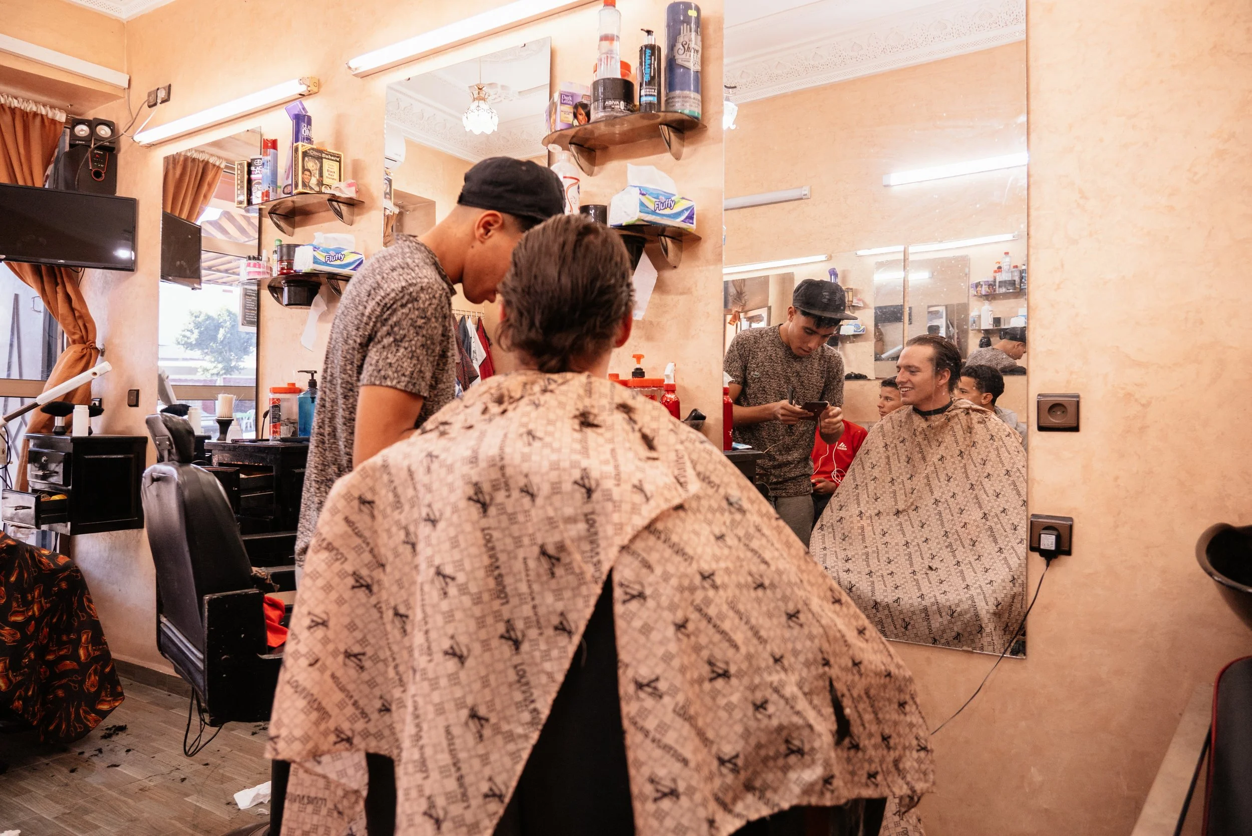 Barbershop scene with two barbers attending to clients, mirrors, mirrors, shelves with hair products, and a wall-mounted TV.