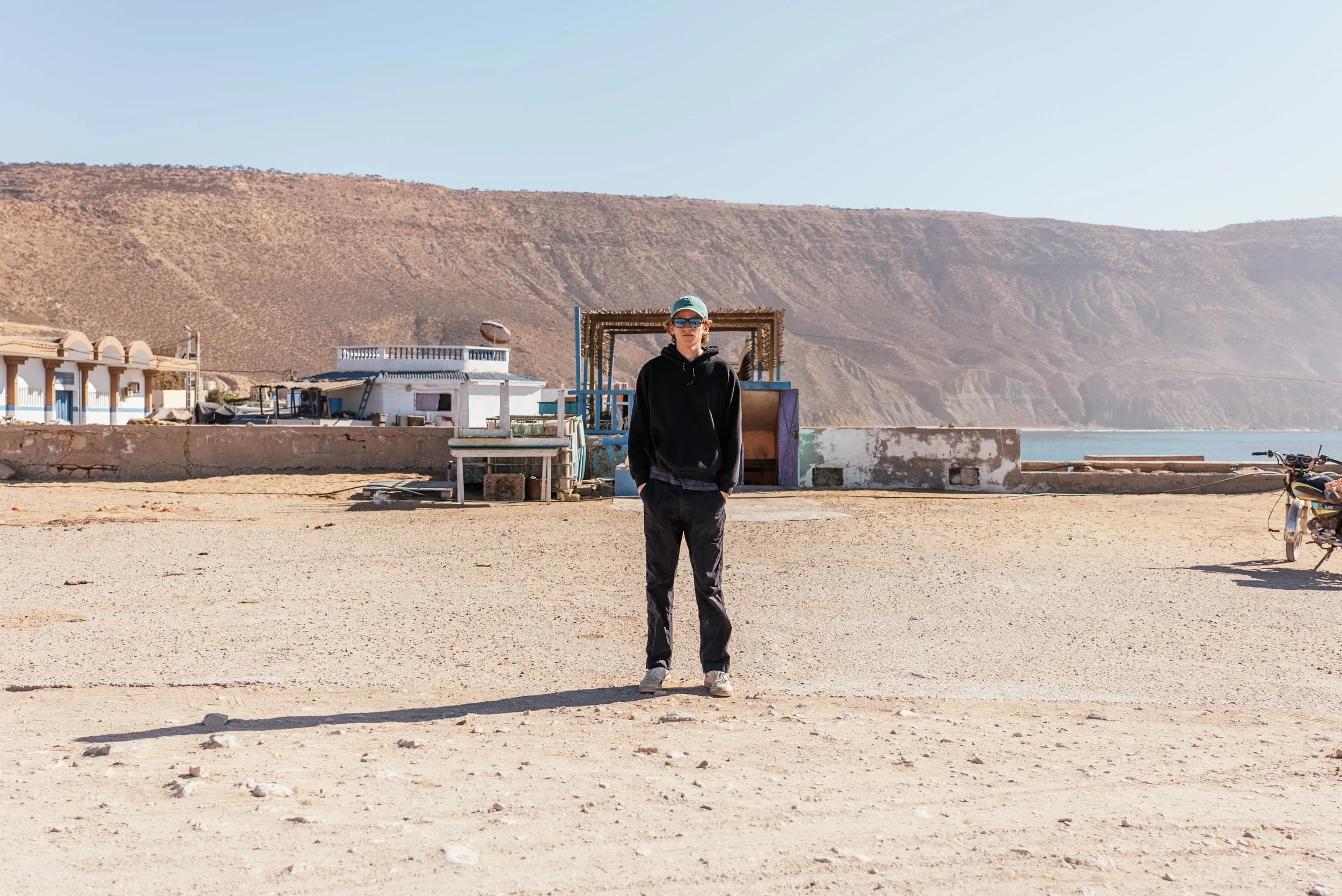 Person standing in a sandy, open area with mountains in the background, wearing dark clothing, sunglasses, and a cap.