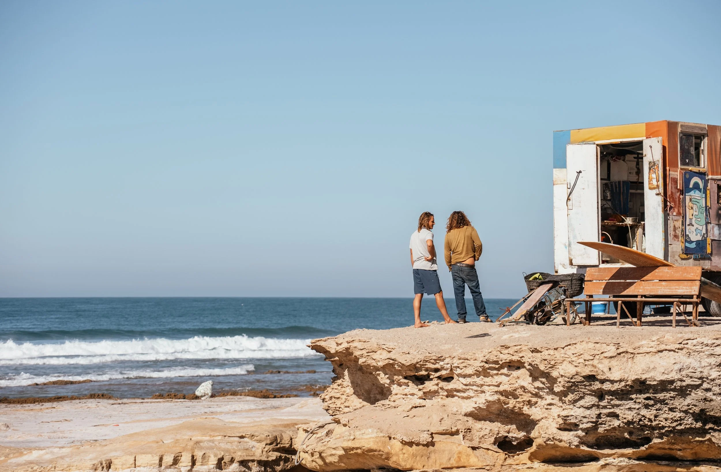 Two people standing on a rocky beach near an artful, colorful beach shack, with the ocean and waves in the background on a clear day.