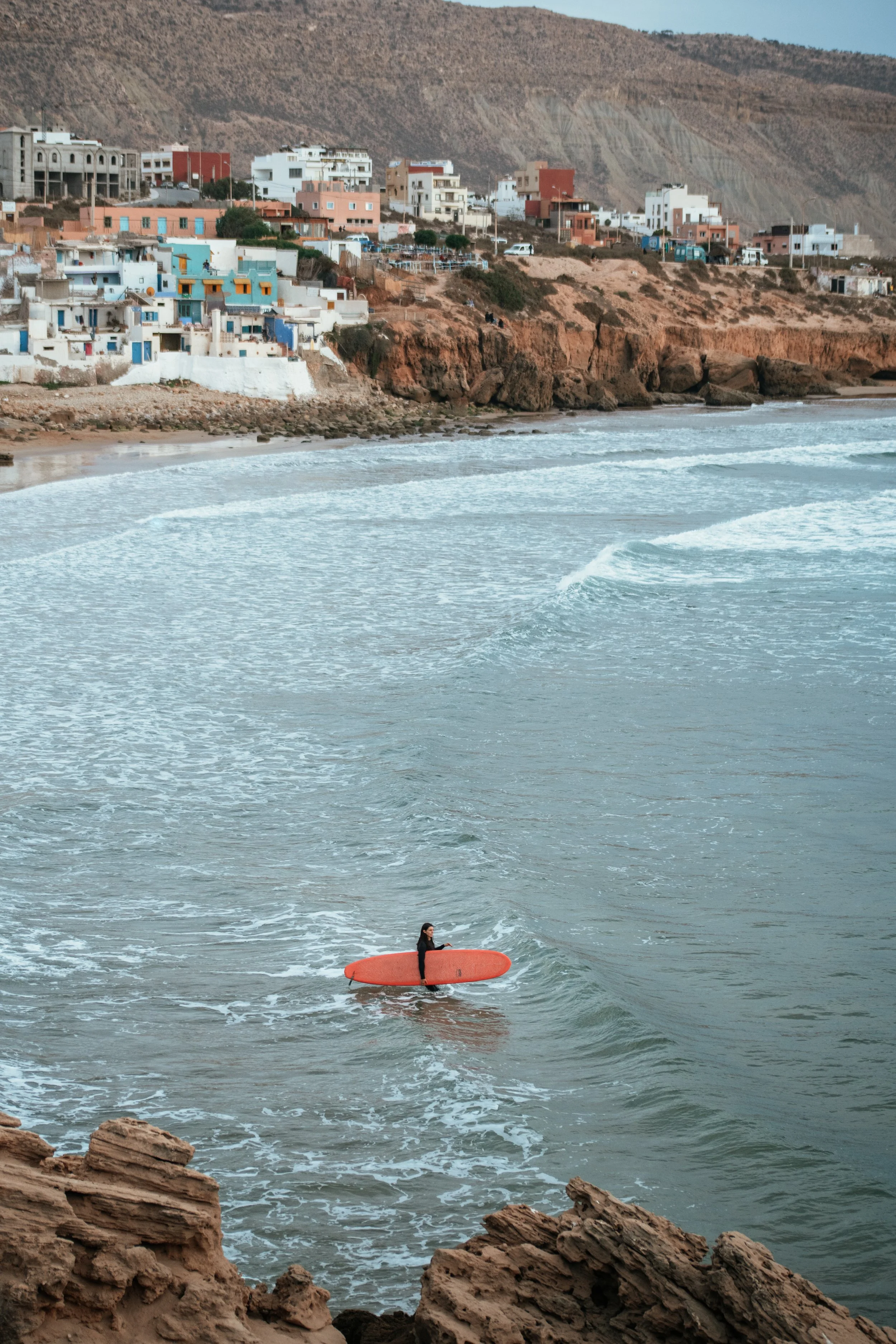 A person holding a surfboard walks in the shallow waters of a beach with rocky cliffs and colorful houses in the background.