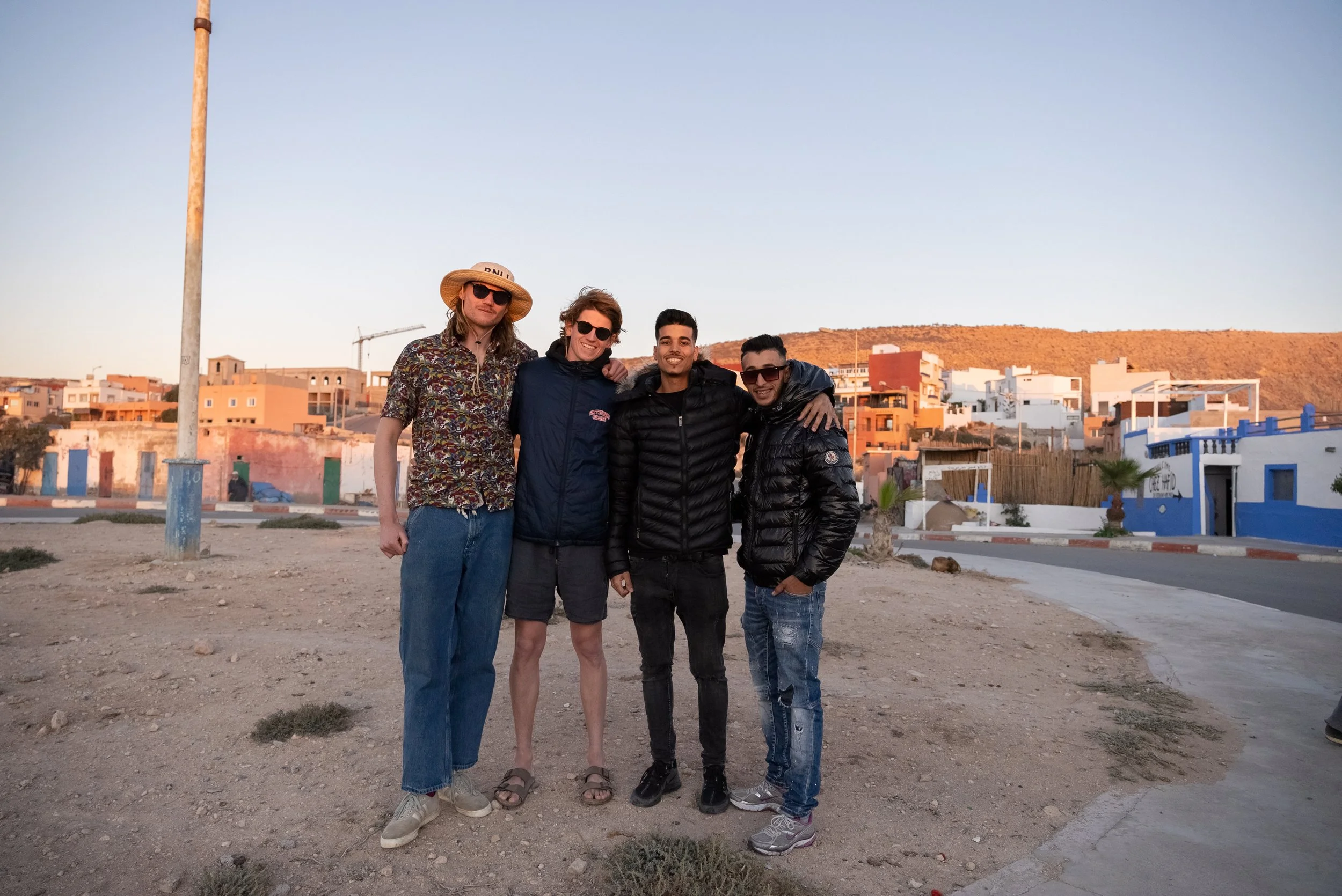 Four men standing together outdoors in a desert-like area with a background of colorful buildings and a hill, during sunset or late afternoon, smiling for the camera.