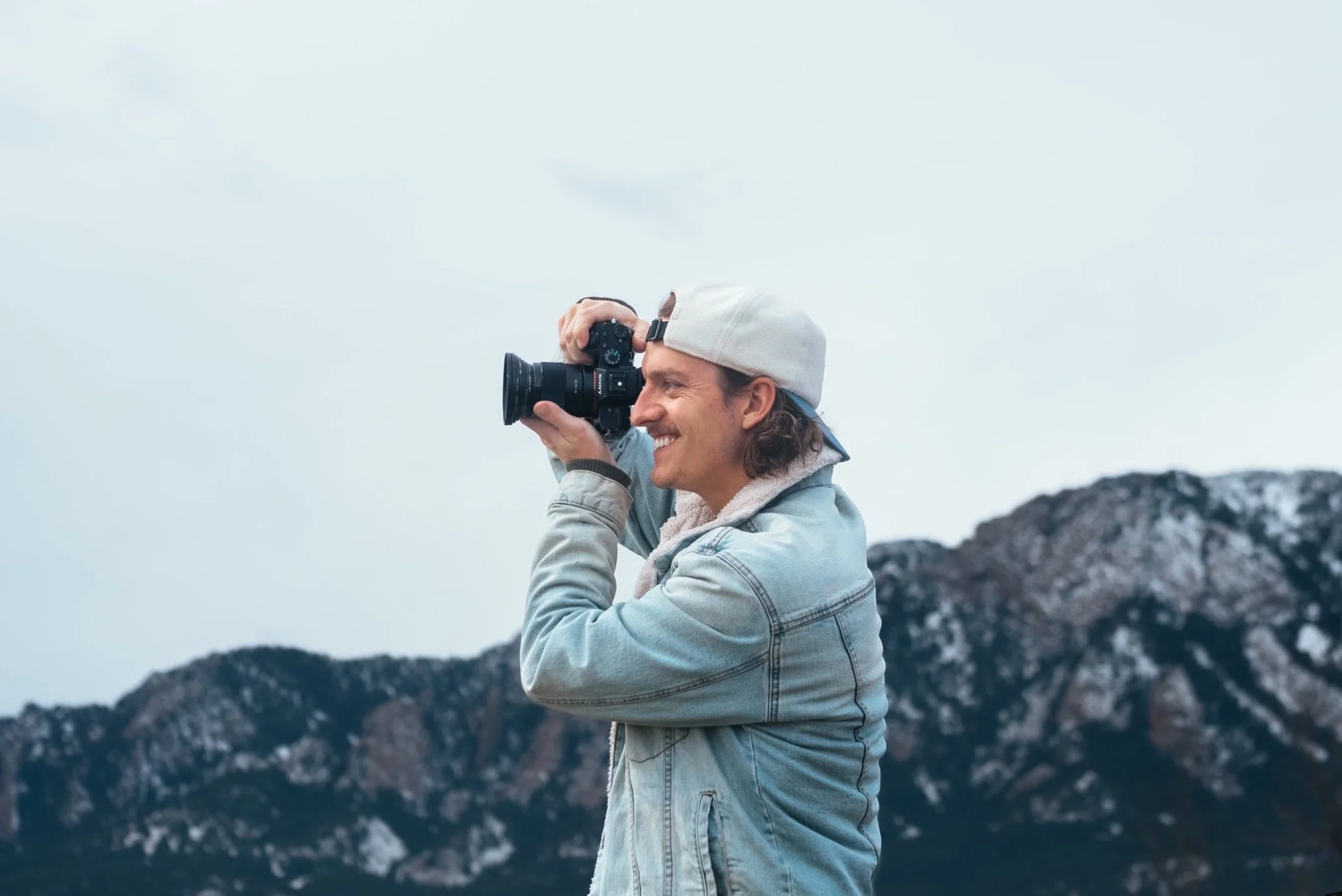 A man wearing a light-colored cap and denim jacket, smiling while looking through a camera, standing outdoors with mountain scenery in the background.