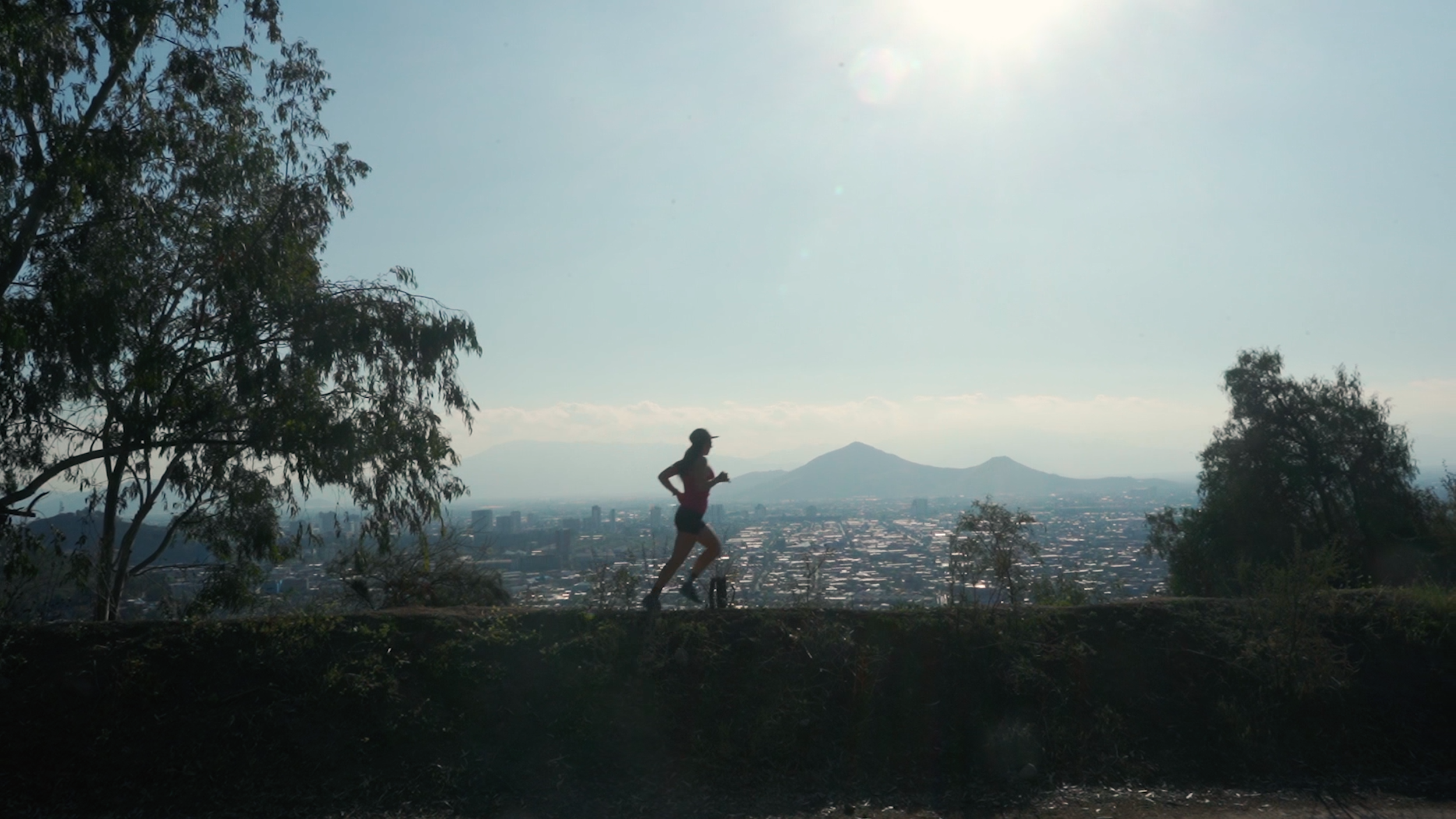 A woman jogging along a trail on a hill, with trees on either side and a city skyline with mountains in the background during the daytime.
