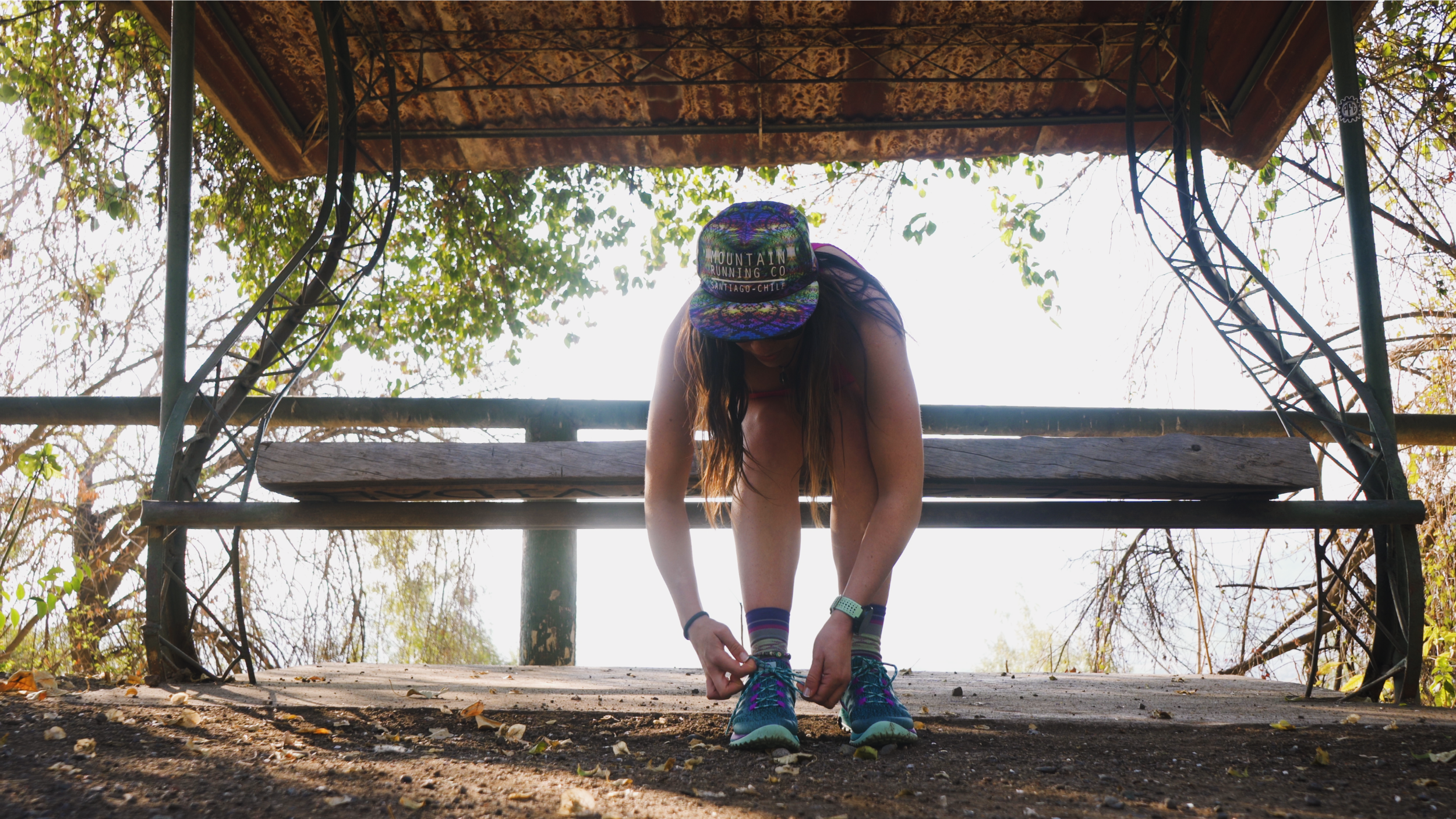 A woman in athletic gear is tying her shoelaces on a wooden platform, wearing a colorful cap and sports shoes, with trees and bright sky in the background.