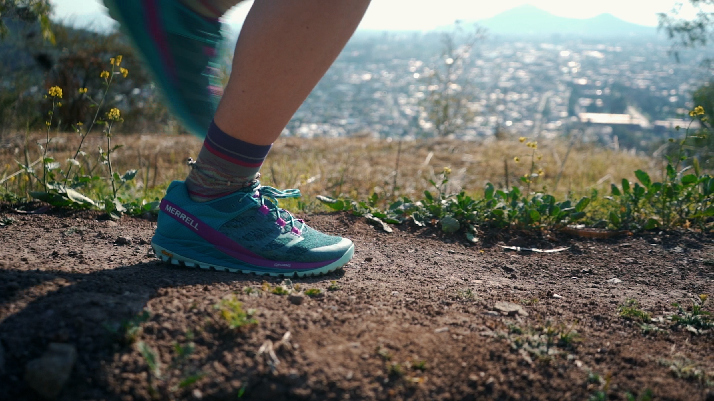 Close-up of a person's foot wearing a bright blue Merrell trail running shoe on a dirt trail, with yellow wildflowers and greenery along the path, and a cityscape in the background.