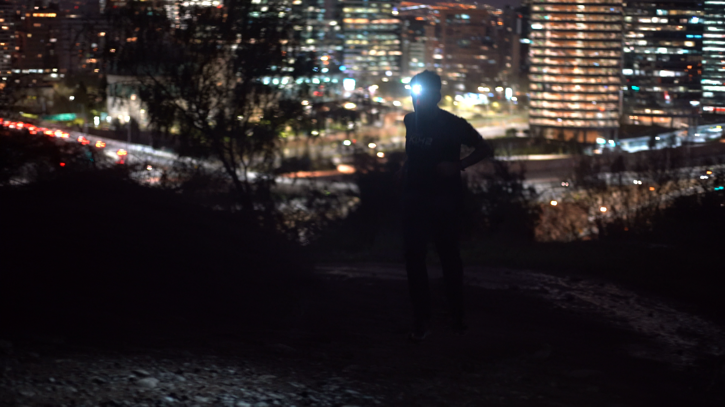 A silhouette of a person standing outdoors at night with a cityscape of lit buildings and traffic in the background.