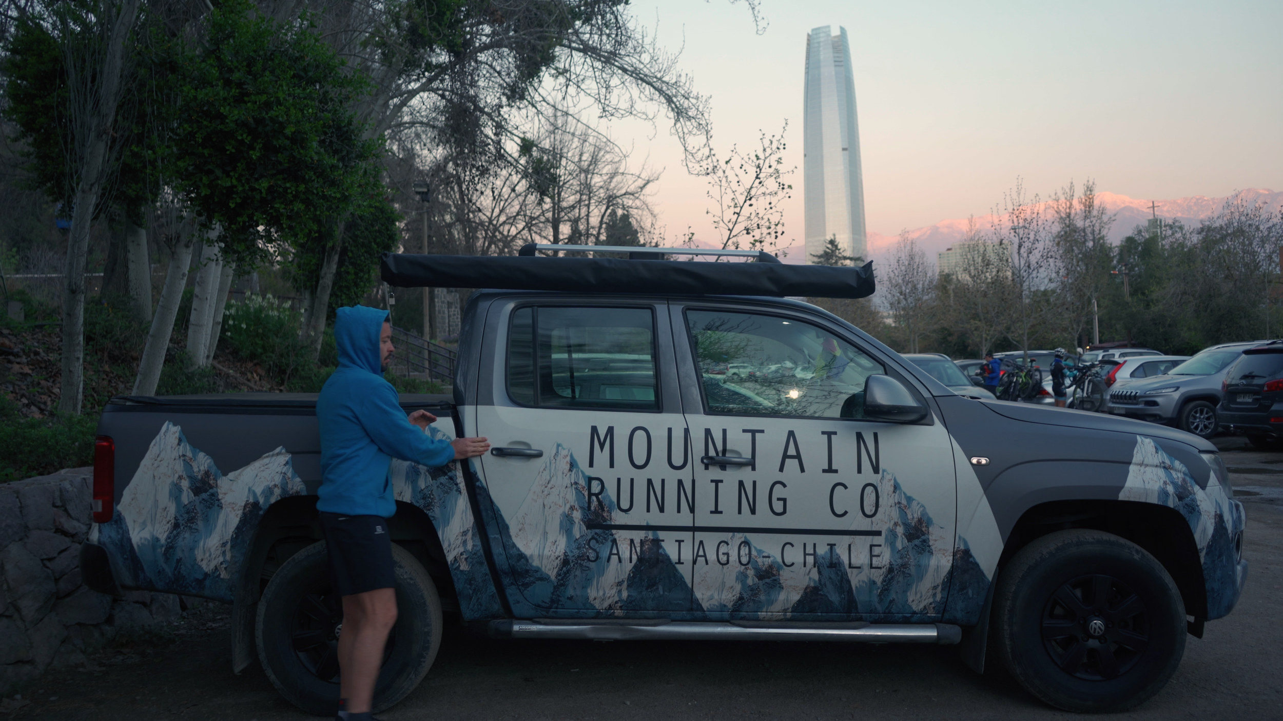 A man in a blue hoodie and shorts standing next to a black and white truck with outdoor mountain graphics, parked in a lot with trees and a tall skyscraper in the background.