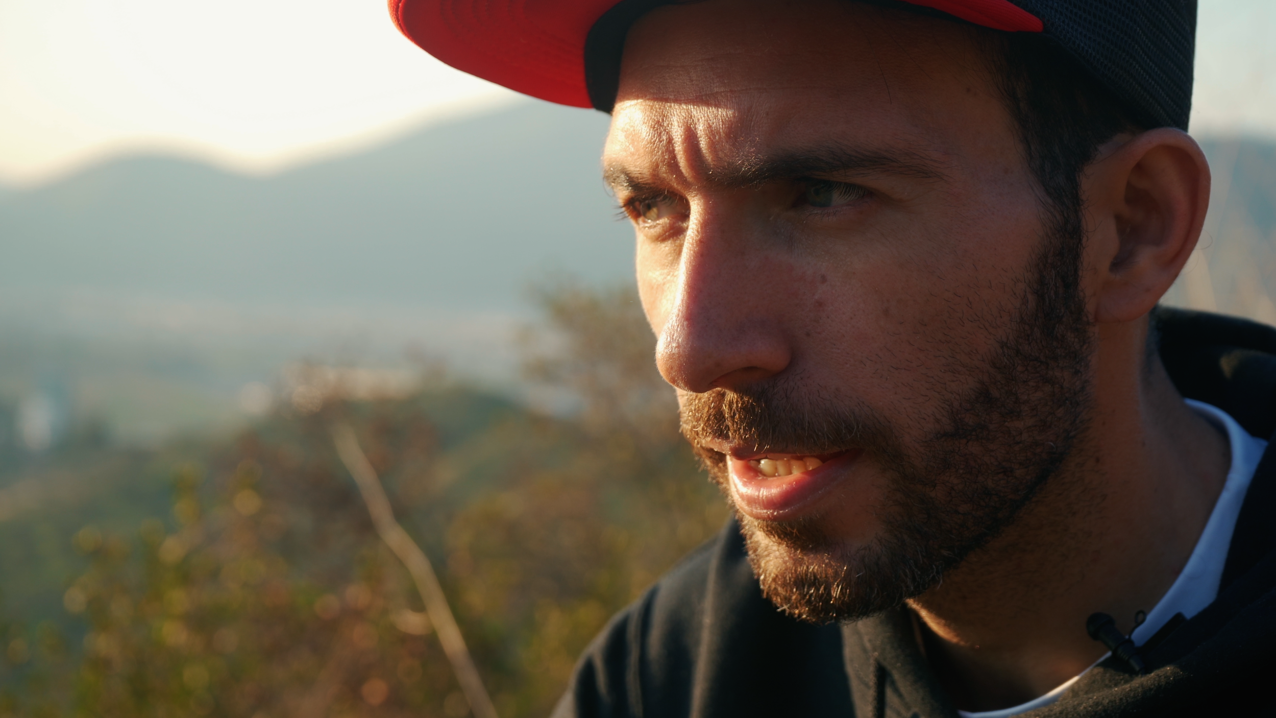 A man with a beard wearing a black and red cap outdoors, looking thoughtful with mountains and trees in the background.