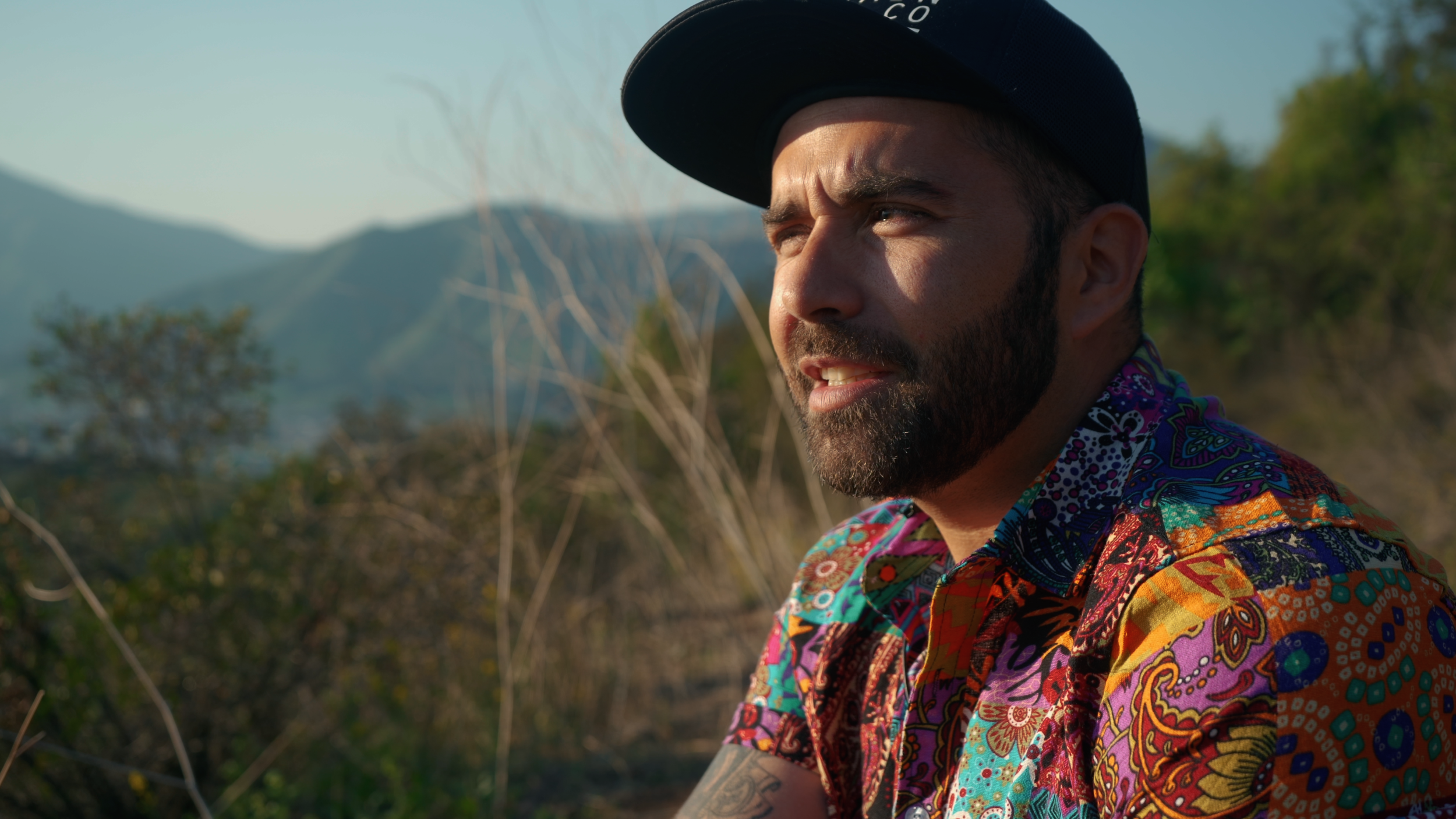 A man with a beard and tattoos wearing a colorful, patterned shirt and a black cap outdoors, with mountains and trees in the background.