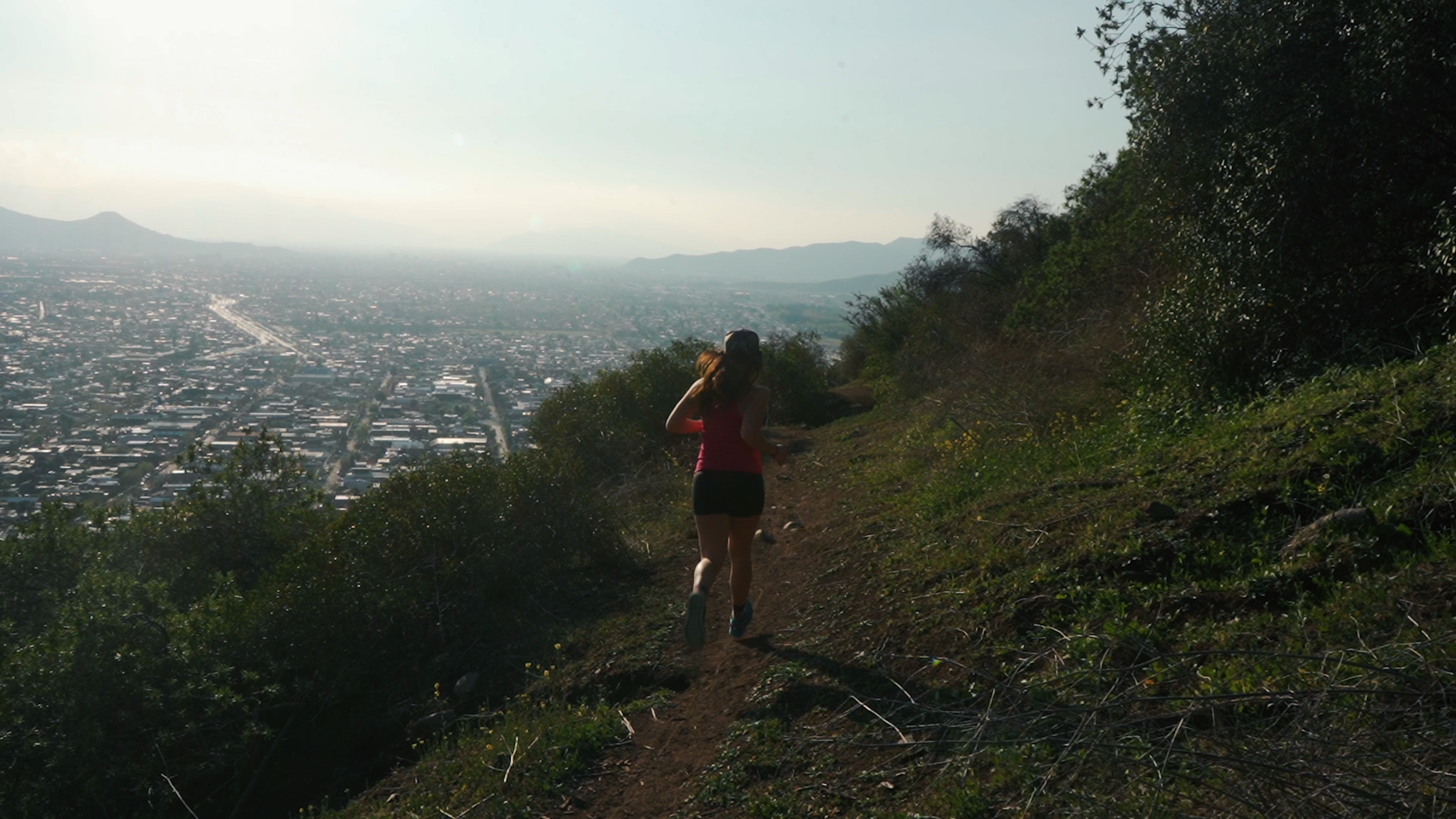 A woman running on a trail on a hillside with a cityscape and mountains in the distance.