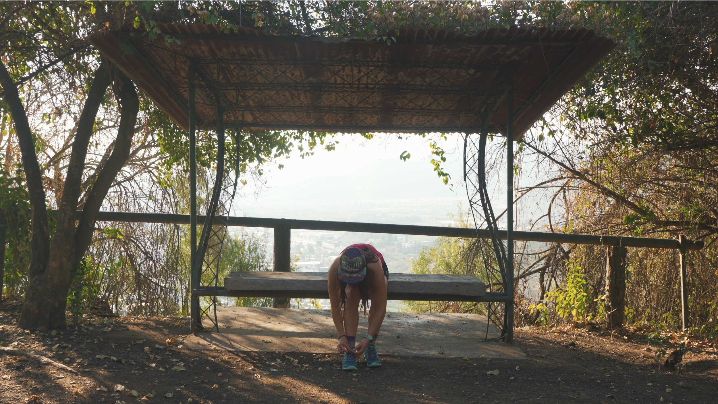 Person tying shoelaces on a bench under a rustic shelter in a wooded area overlooking a cityscape and distant mountains.