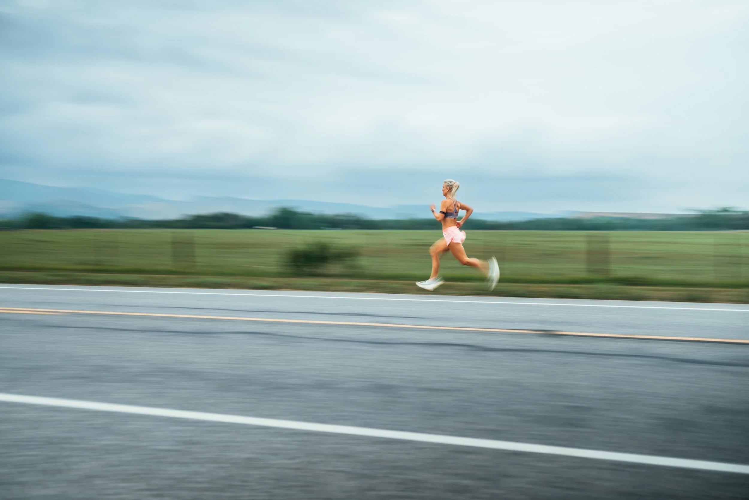 A woman running on a road in a rural area with green fields and overcast sky in the background.