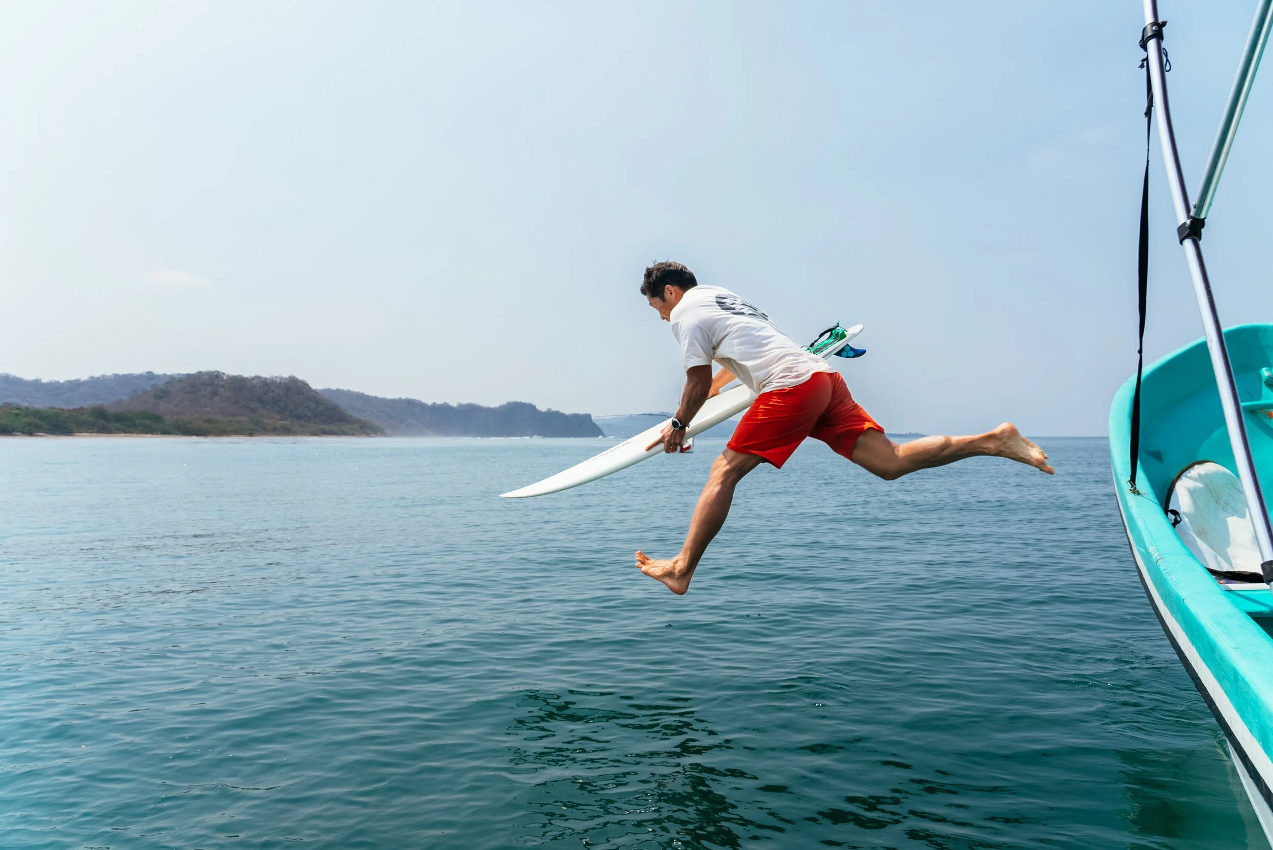Man jumping off boat into water with surfboard in hand, wearing red shorts and white t-shirt, near a small boat on a clear day.