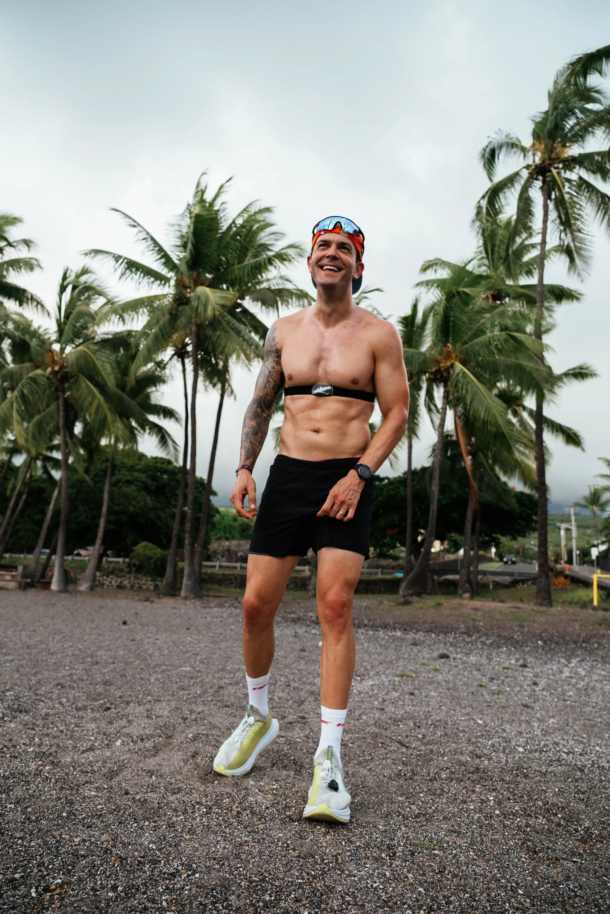 A man running outdoors among palm trees, shirtless, wearing black shorts, running shoes, white socks, and a headband with goggles on his forehead, smiling.