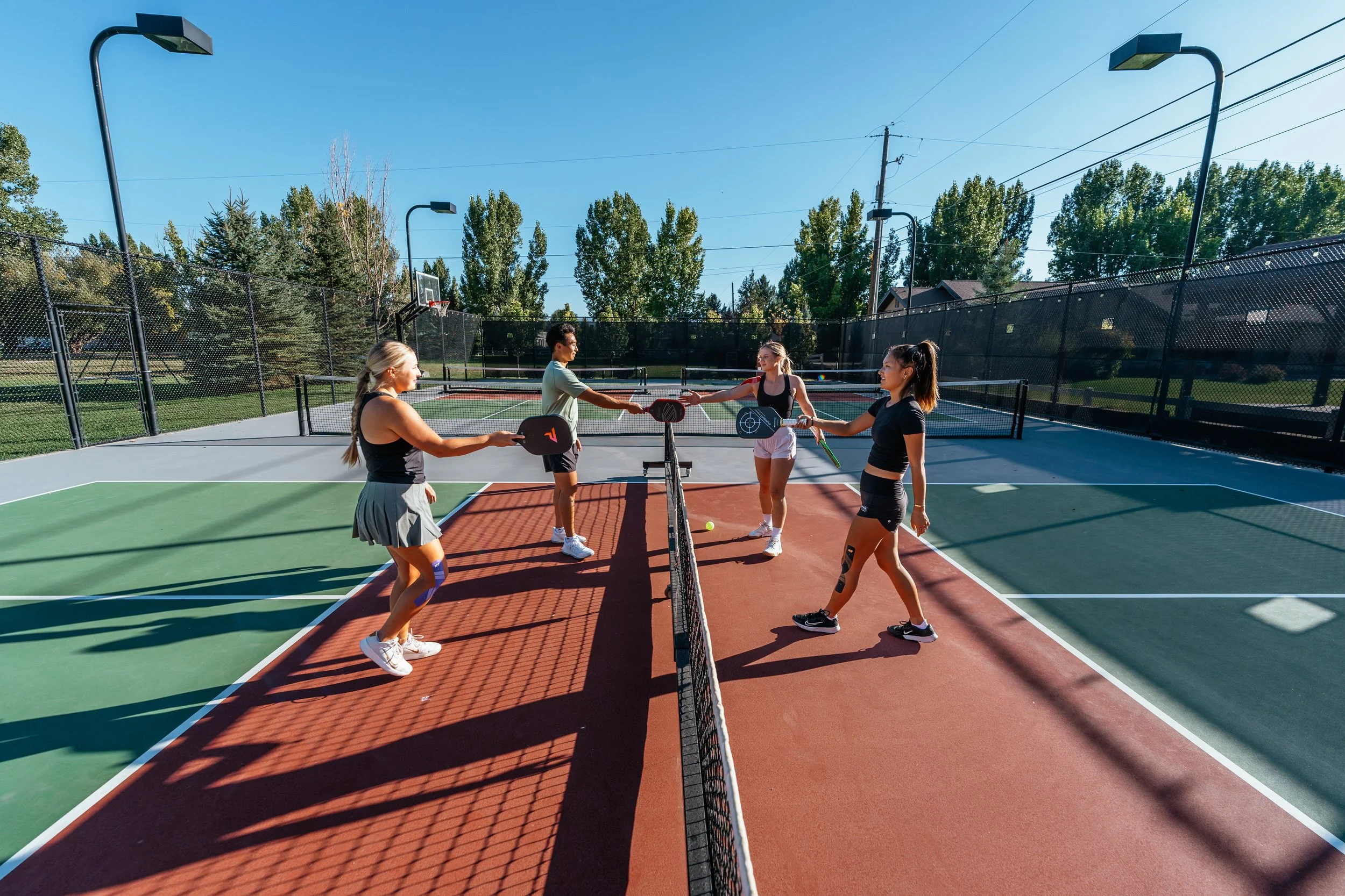 Four women and one man playing pickleball on outdoor courts, with trees and a clear blue sky in the background.