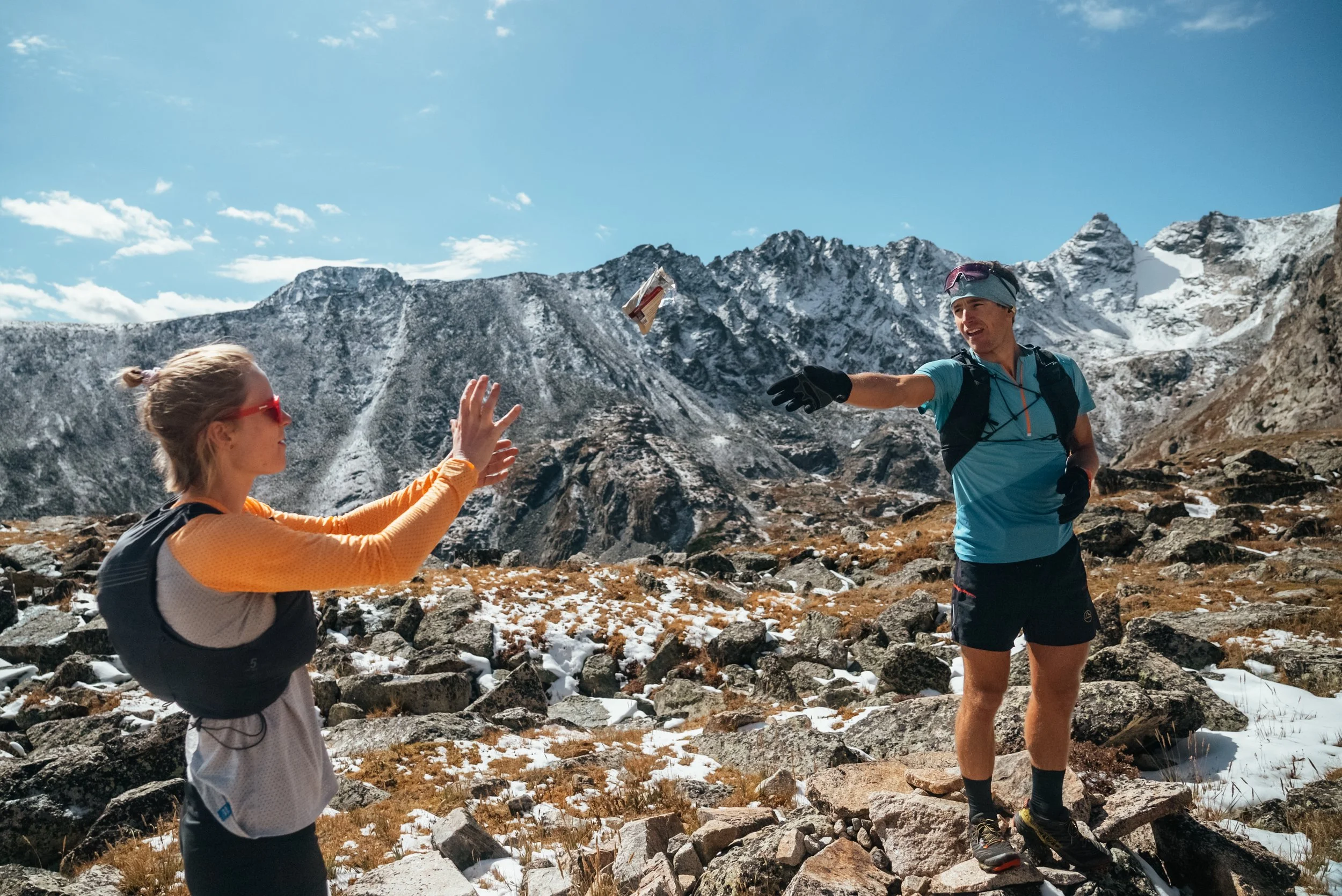 A woman in sunglasses and outdoor gear reaching out to catch a snack from a man in hiking attire, set against snowy mountain peaks and rocky terrain.