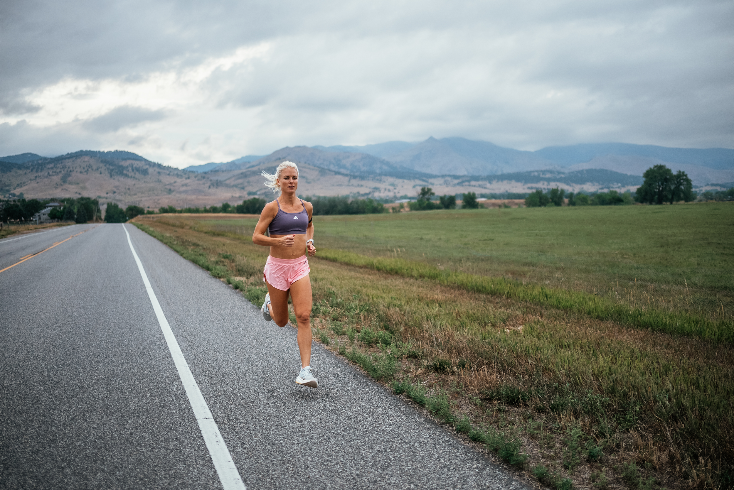 A woman running on the side of a rural road in a landscape with mountains and cloudy sky.