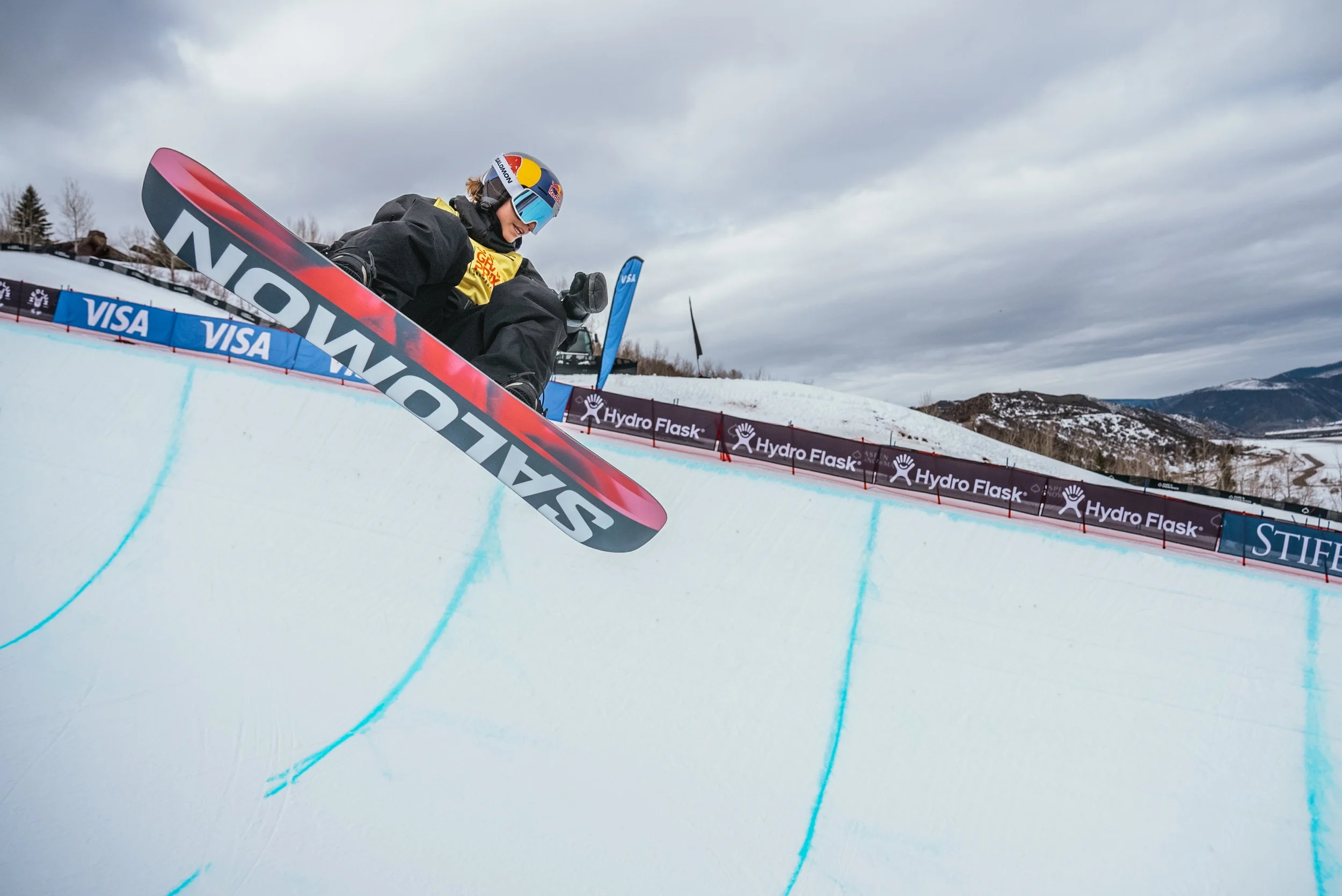 A snowboarder wearing a helmet and goggles jumps off a ramp during a race, with snow-covered mountains and cloudy sky in the background.